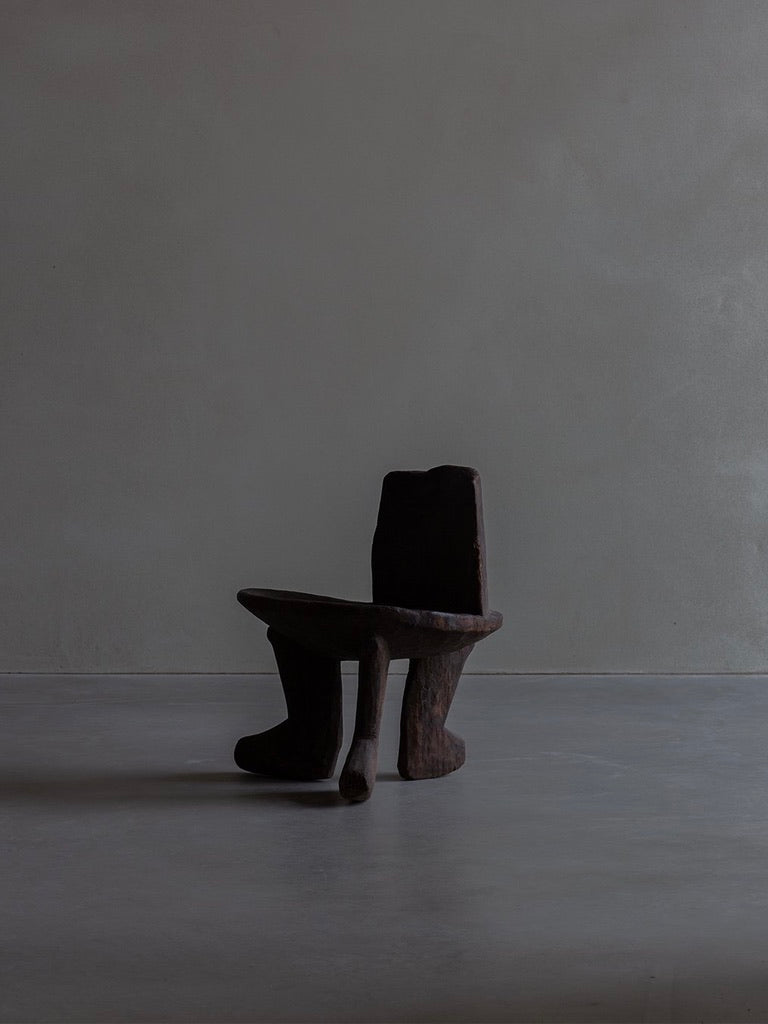 The African Stool by Bicci de' Medici, with its unique dark wooden design reminiscent of primitive art, stands on a smooth light gray floor against a plain gray wall, creating a minimalist and artistic scene that reflects African craftsmanship.