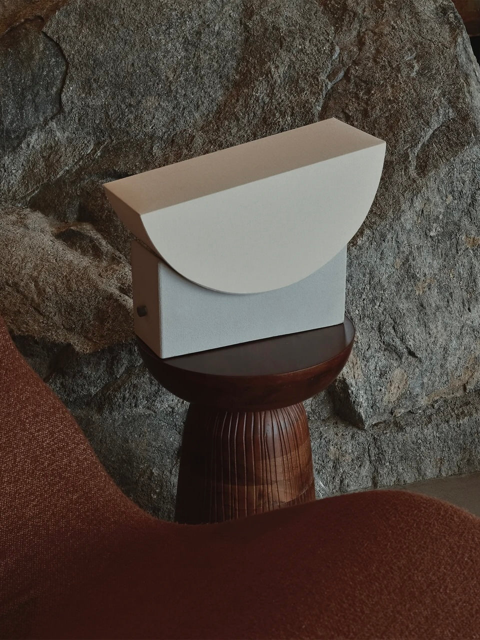 Introducing the "Reminder" Table Lamp by Enkei Lights AB, featuring a sleek and modern aesthetic with geometric shapes. The lamp boasts a curved white top alongside a rectangular gray base and is elegantly displayed on a wooden stand. In the background, rocky elements add texture, while brown fabric edges frame the scene in the foreground.