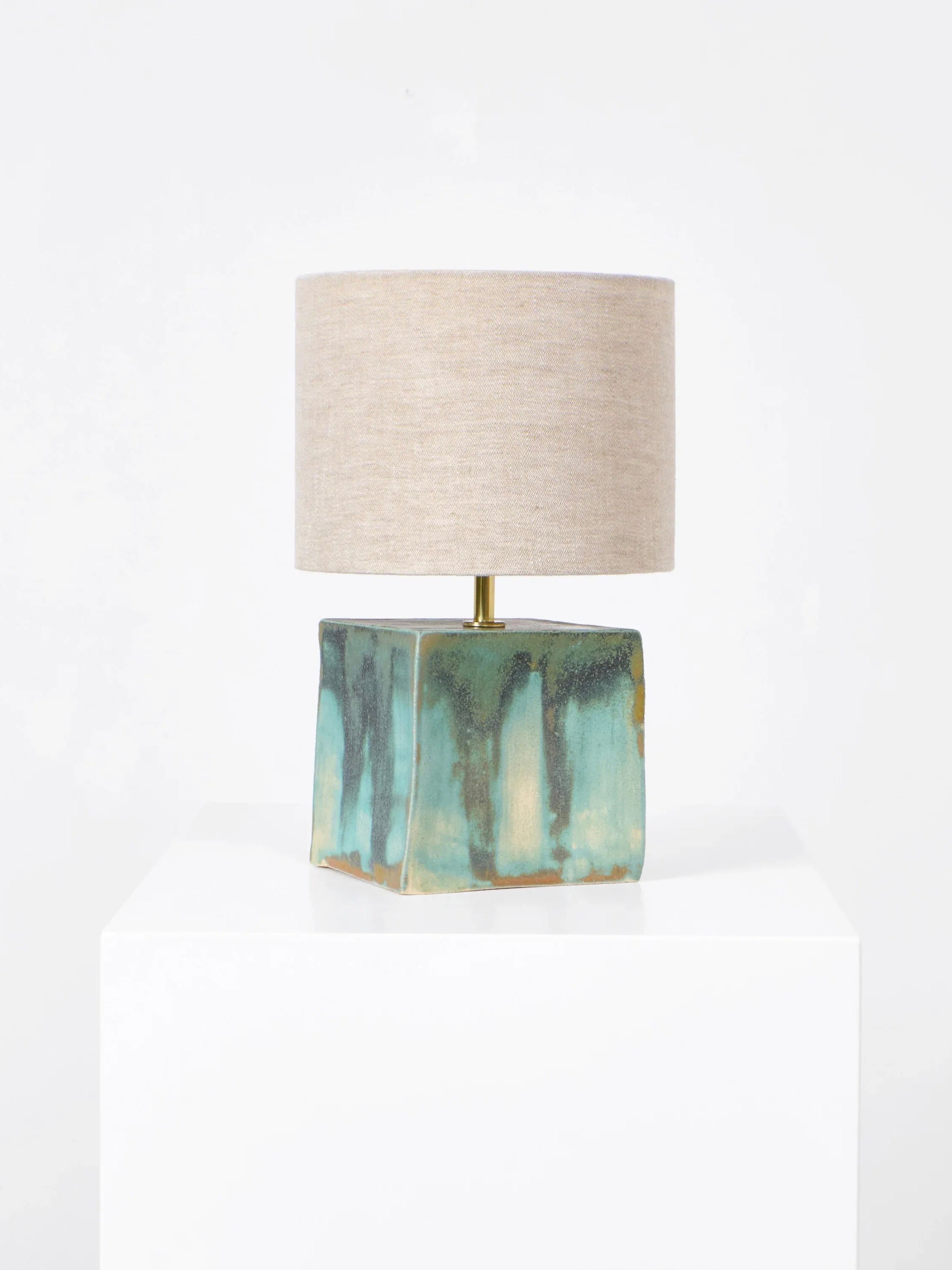 The Arouca Table Light by Project 213A features a handmade rectangular ceramic base in green and brown glaze, topped with a cylindrical beige fabric shade. It sits elegantly on a white pedestal against a plain white background.