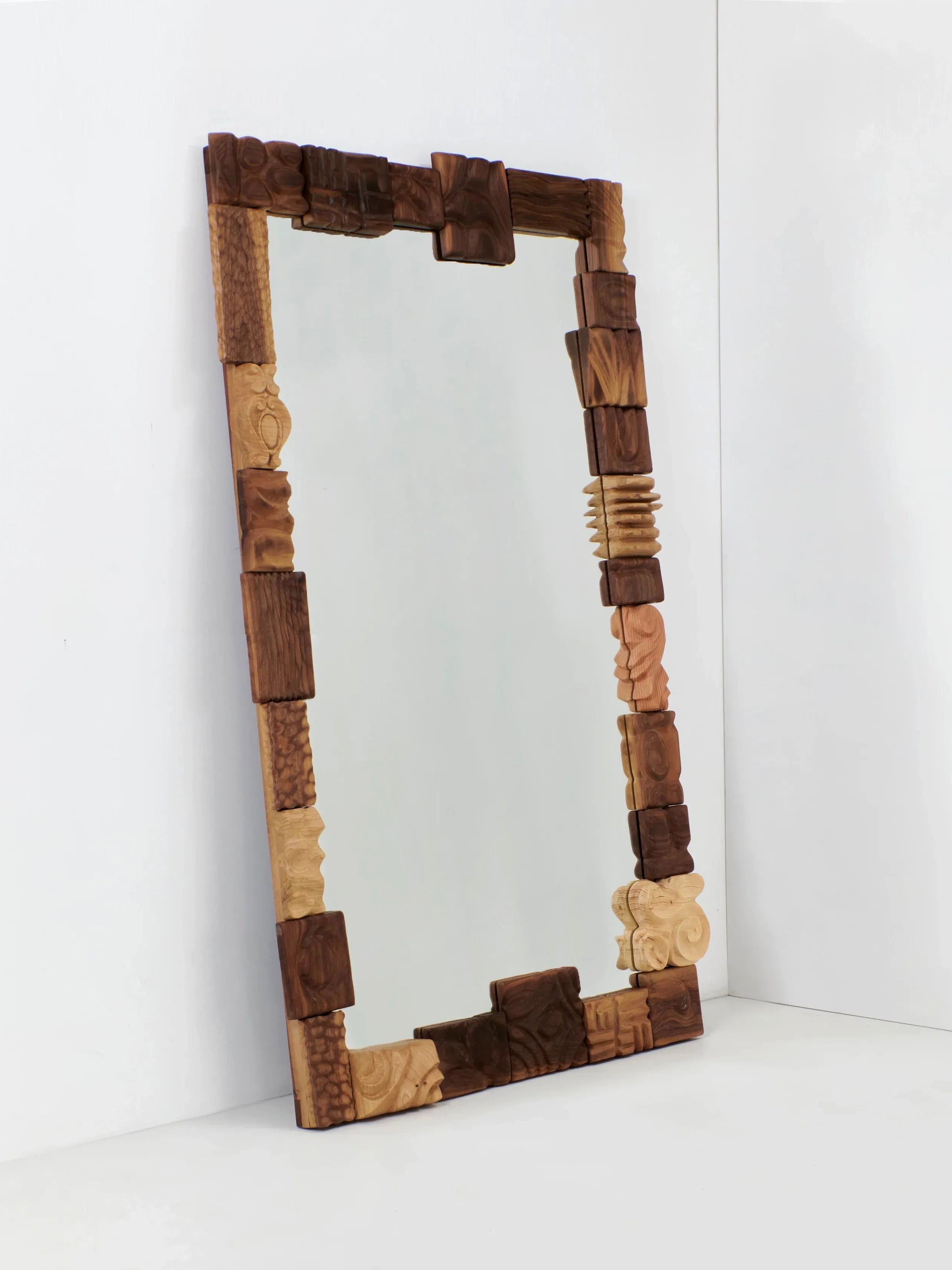 The Catarina Mirror by Project 213A, a large wall-leaning piece, stands elegantly against a white wall. Its frame boasts hand-carved ornamental wood blocks in an array of shapes and shades, imparting a distinct and artistic flair to the mirror.