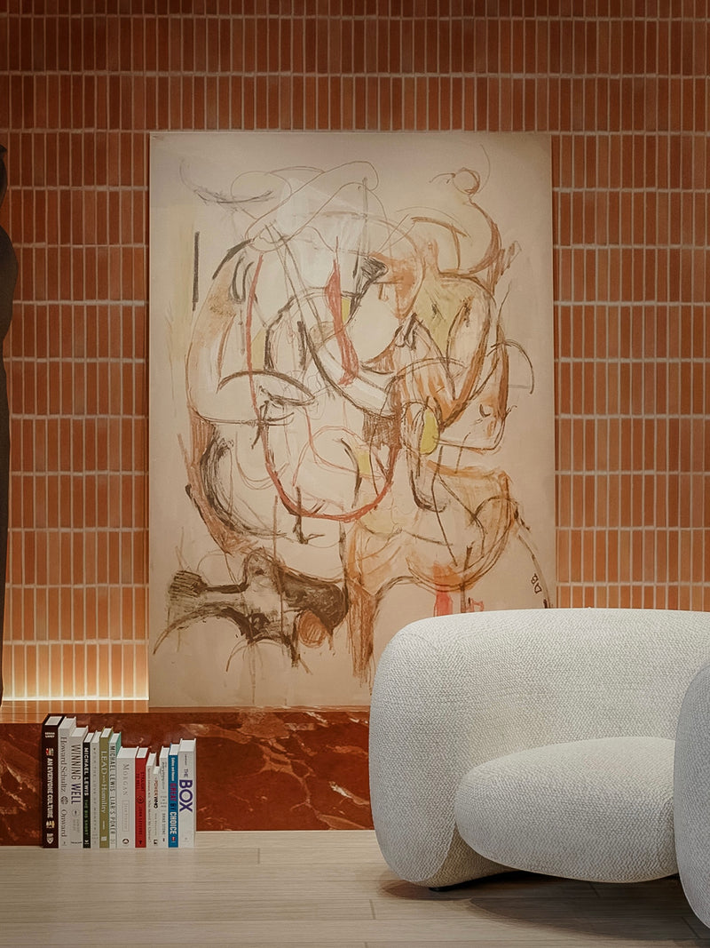 Memory of the Body," a Dovile Bernotaite painting, exhibits abstract swirls and dynamic lines in earthy tones on a textured vertical-patterned wall. A plush white rounded chair is placed by books on a red marble surface in the foreground.