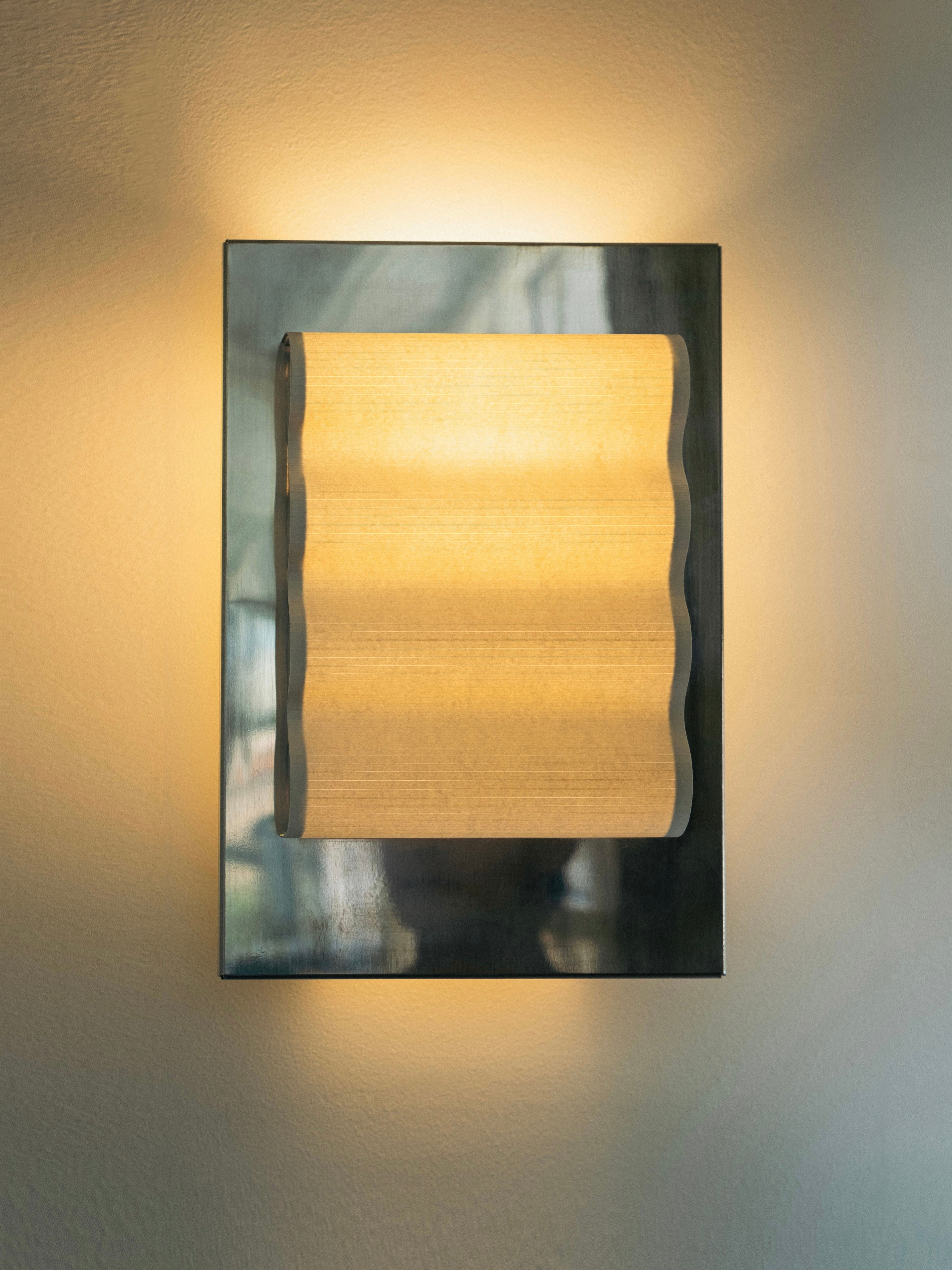 Introducing the Wall Light Frame Miroir S by Violaine d'Harcourt—a sleek, wall-mounted light fixture that emits a soft, warm glow. This minimalist design features a rectangular metallic base with a wave-patterned translucent cover that evenly diffuses the light, creating exquisite ambient illumination against the wall.