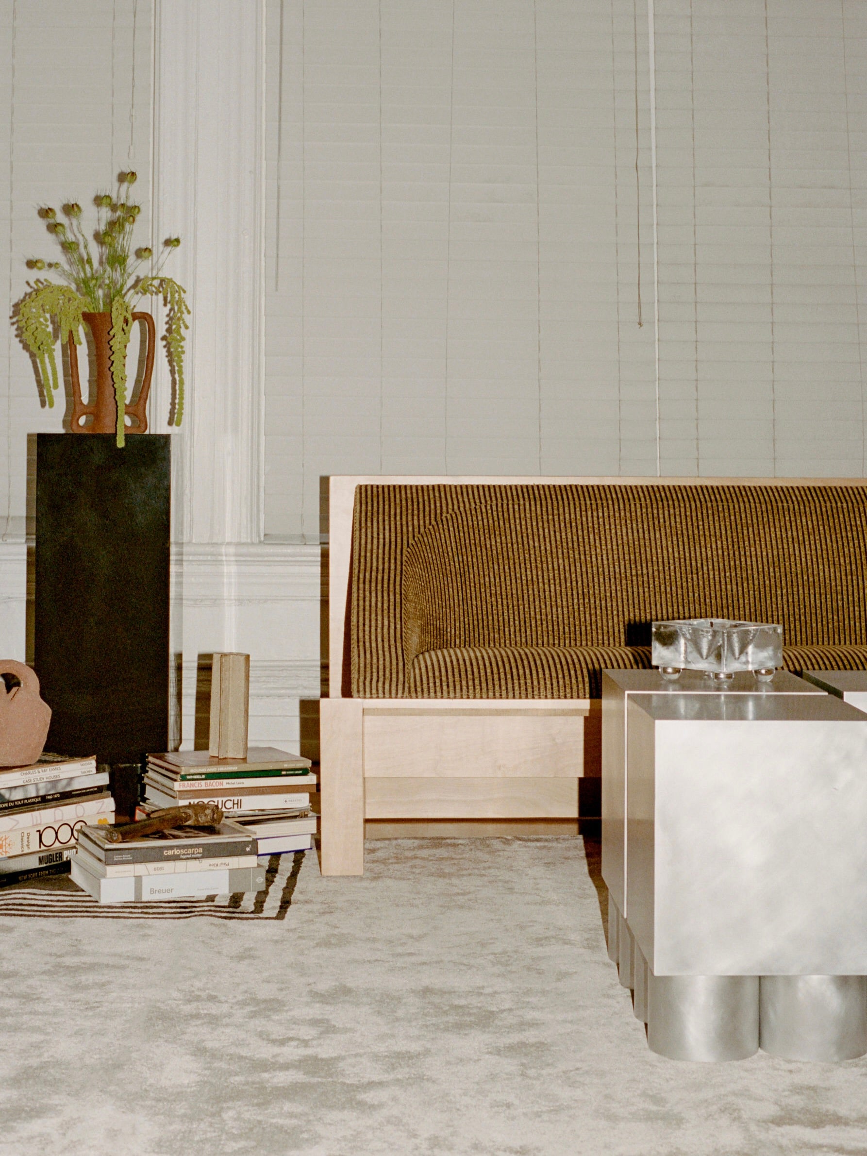 A cozy living room showcases the Known Work 'Perceptions' Sofa Bench with a plush striped upholstery, a metal table with a patterned top, and glasses. A tall vase on a black pedestal reflects minimalist values, while stacked books and blinds covering large windows create an inviting ambiance.