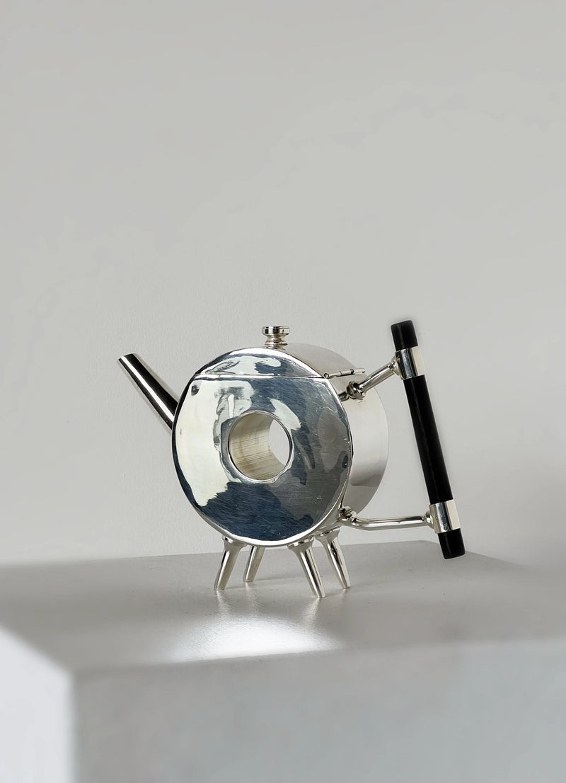 The Christopher Dresser Ornate Round Teapot by Porter Studio features a modern circular silver-plated design with a black handle and four angled legs, displayed on a white surface against a plain light gray background.