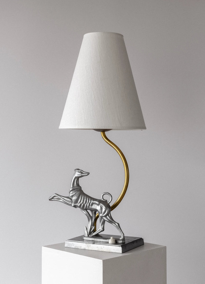 The Spigel Art Deco Greyhound Table Lamp (1930s) features a white conical shade, curved gold stem, and silver greyhound base, shown on a white pedestal against a plain light gray background.