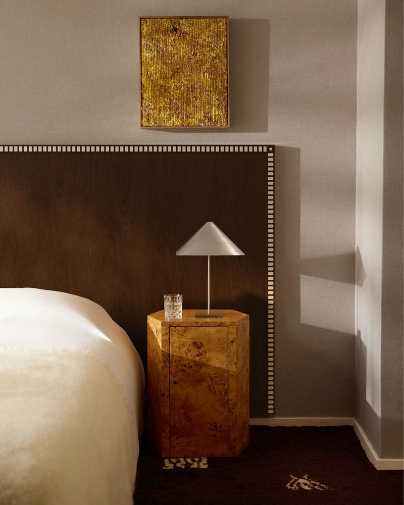 A modern bedroom features a wooden headboard, soft lighting, textured gold artwork, and Lemon's Pair of Park Night Stands in Poplar Burl—Italian design pieces that hold a glass of water and a metallic lamp, showcasing exceptional detailing throughout.