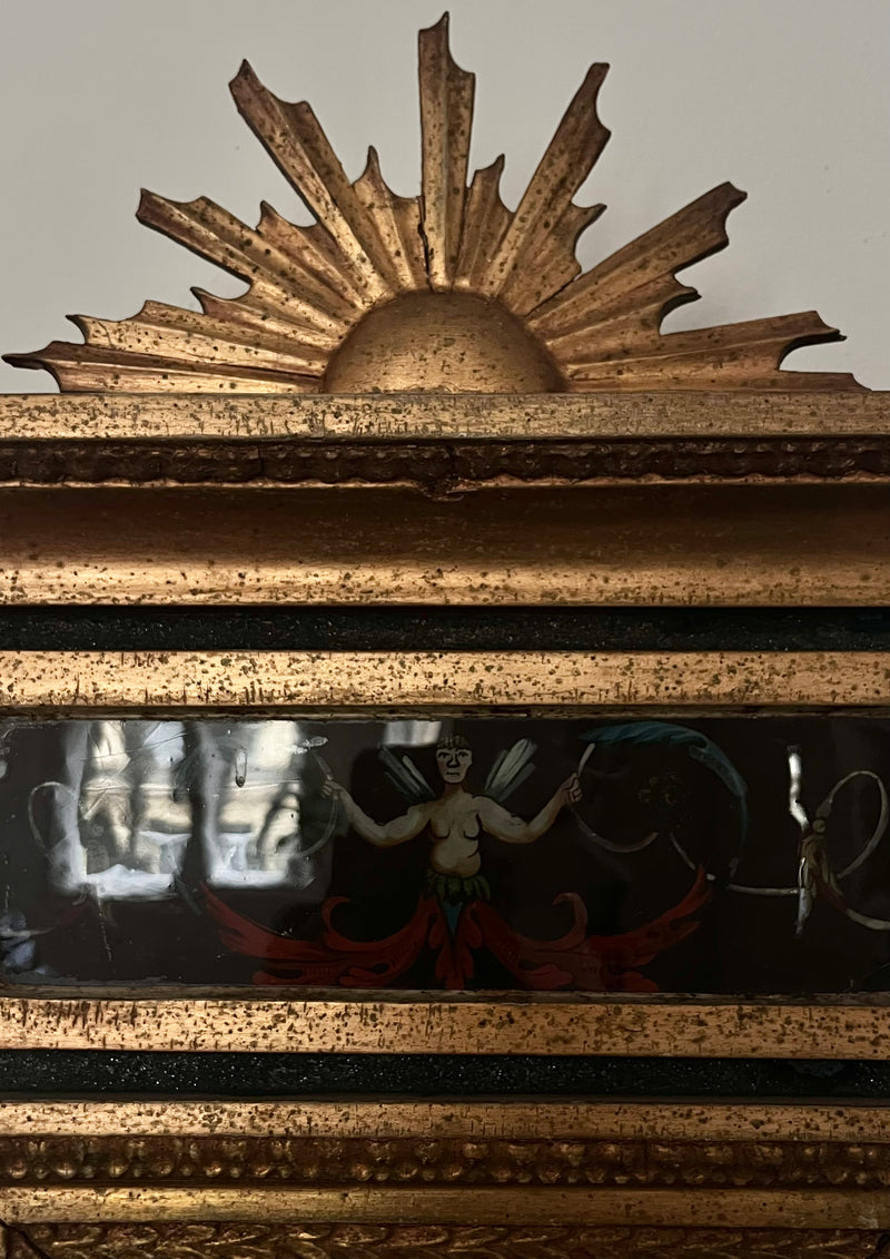 The Médecine Antique Gilded Mirror showcases an ornate, sunburst-topped frame and a painted panel below featuring a winged figure with outstretched arms, set amid dark and red hues.