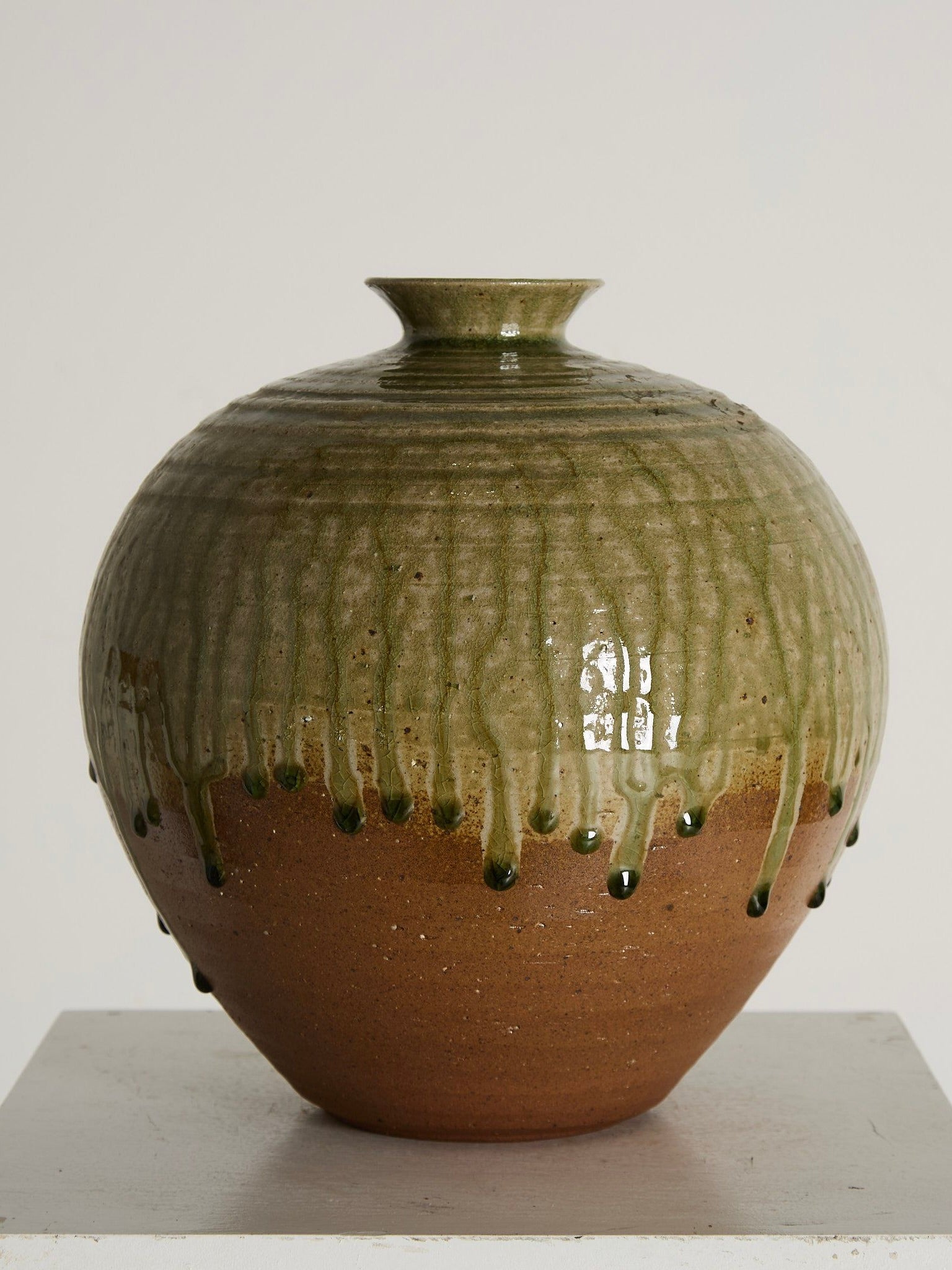 The Drip Glazed Vase by Ancien et Jolie, from Hokkaido, Japan, highlights mid-century elegance with its shiny green drip glaze over a reddish-brown base. Set on a white pedestal against a plain background, it exudes timeless sophistication.