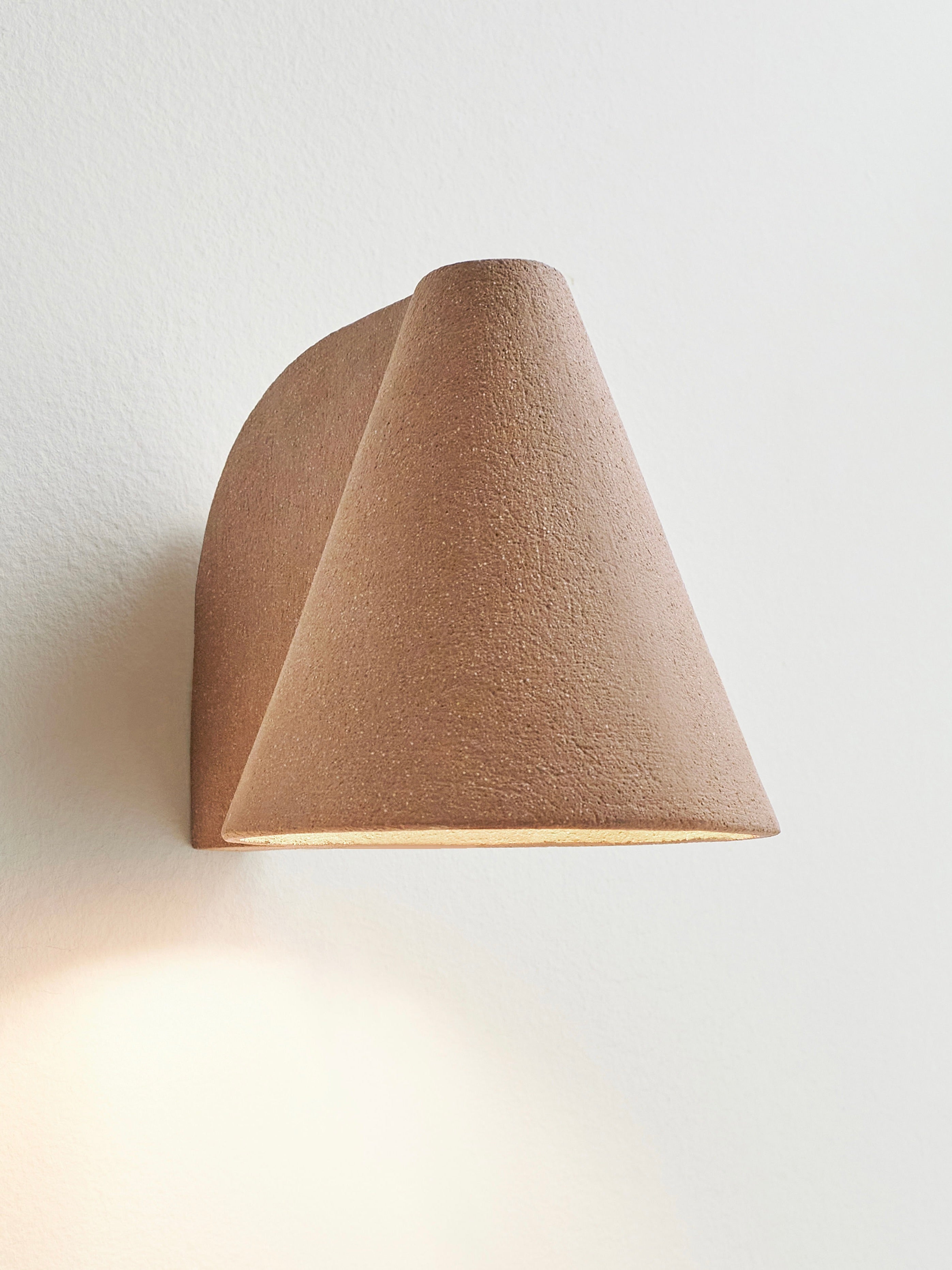 A Mariza Galani 'Esoteric' Almond Brown Wall Light, handcrafted in a minimalist cone shape with an earthy finish, is mounted on a plain white wall. It casts a soft glow below, creating a warm and inviting atmosphere.