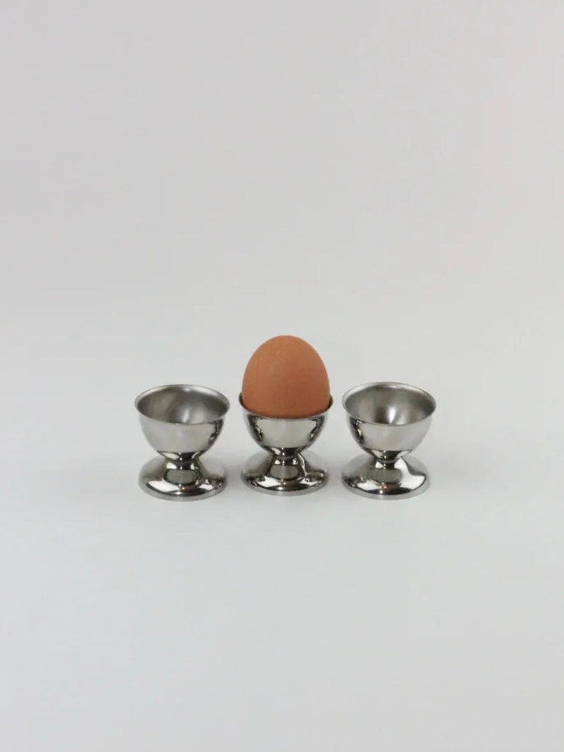 A single brown egg rests in the middle of three shiny stainless steel "Six Inox Egg Cups" from Boga Avante Shop, neatly arranged in a row on a plain white background. One egg cup holds the egg, while the other two remain empty. These elegant breakfast pieces from Boga Avante Shop showcase both durability and strength.