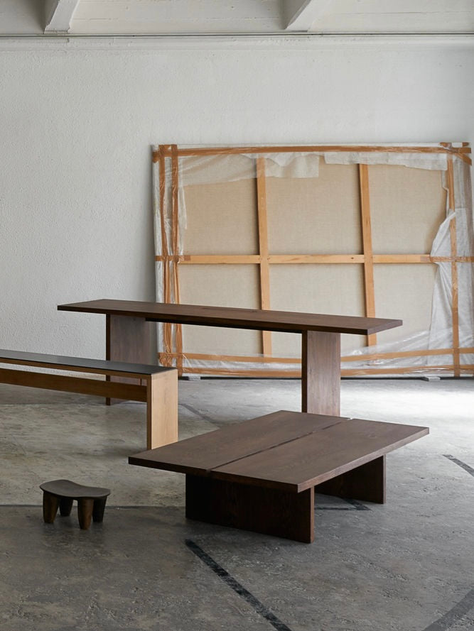 The minimalist room boasts a Tanizaki Coffee Table by ASERIES OF OBSTACLES, SL, set on a concrete floor with a modern dark wood bench. A small wooden stool invites seating near the ground, while a large canvas leans against the white wall in the background.