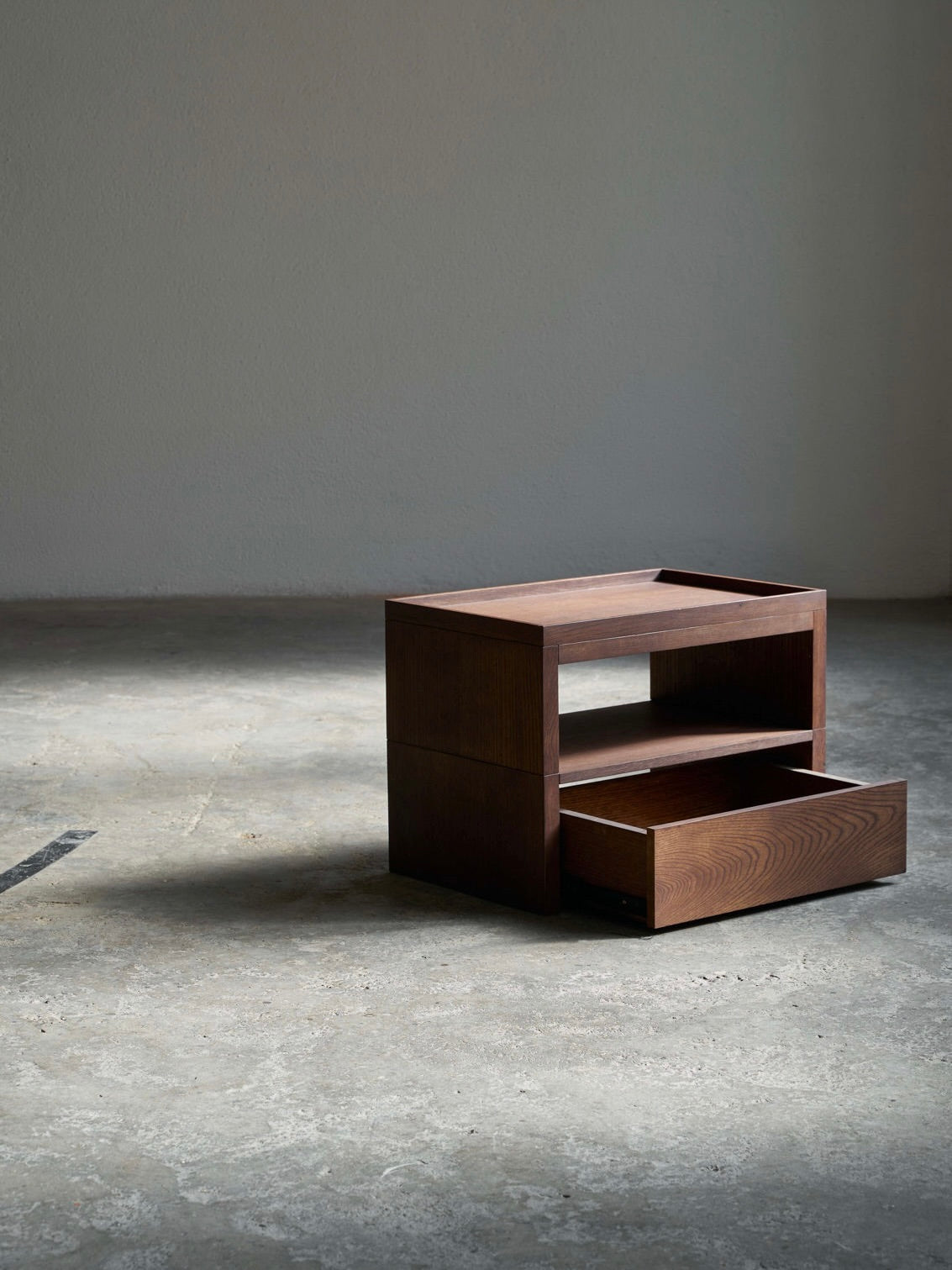 The ASERIES OF OBSTACLES, SL Eileen Bedside Table + Tray features a slightly open tray drawer on a textured concrete floor, set against a plain wall. Soft lighting highlights the wood grain while casting gentle shadows.