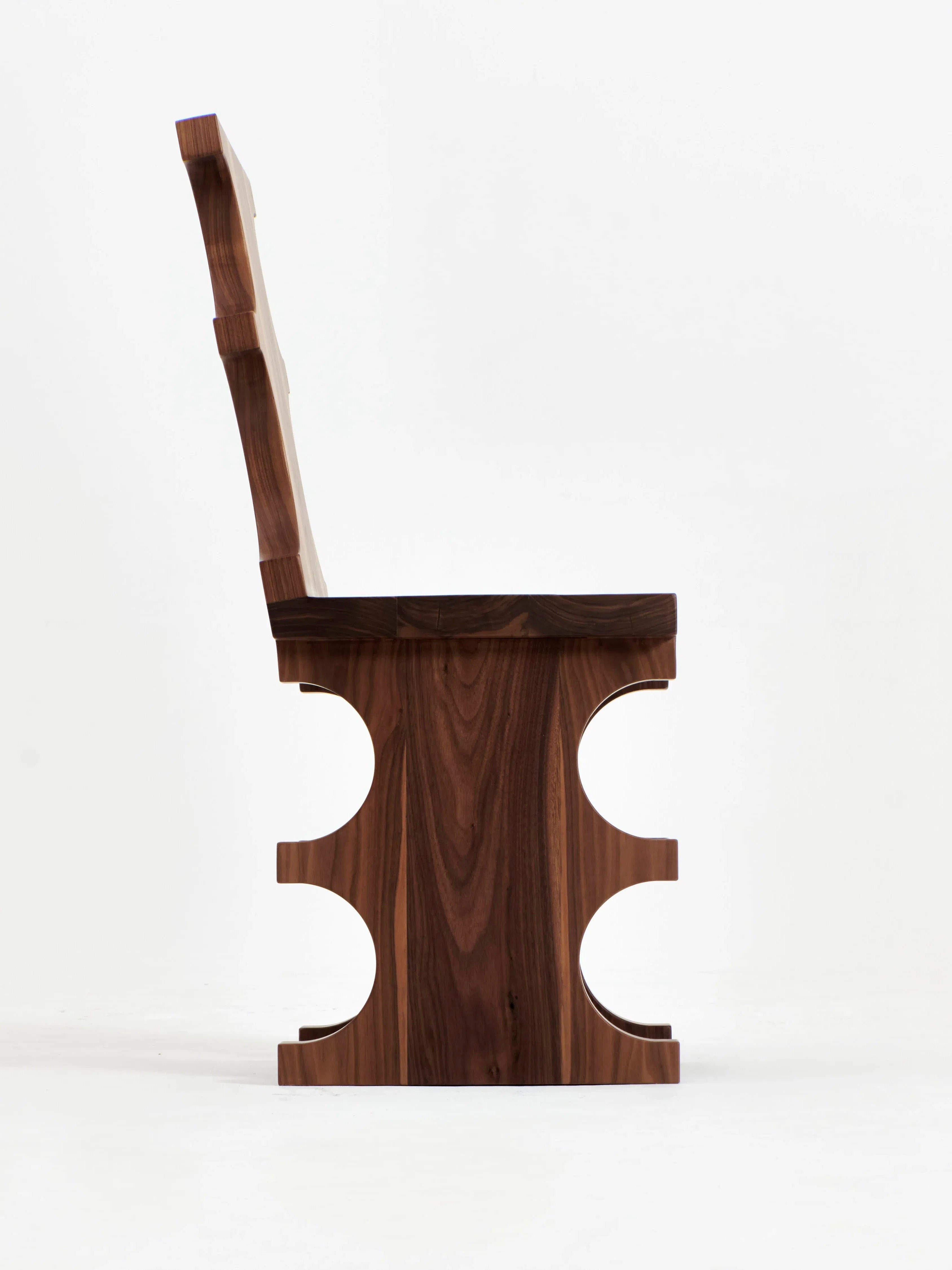 A side view of the Portugal Chair No 7 by Project 213A showcases its unique, sculptural design. The chair features a solid backrest and seat with a cut-out pattern in the center of the legs, creating a modern and artistic aesthetic. Crafted by skilled artisans, the solid walnut wood boasts a smooth, polished finish.