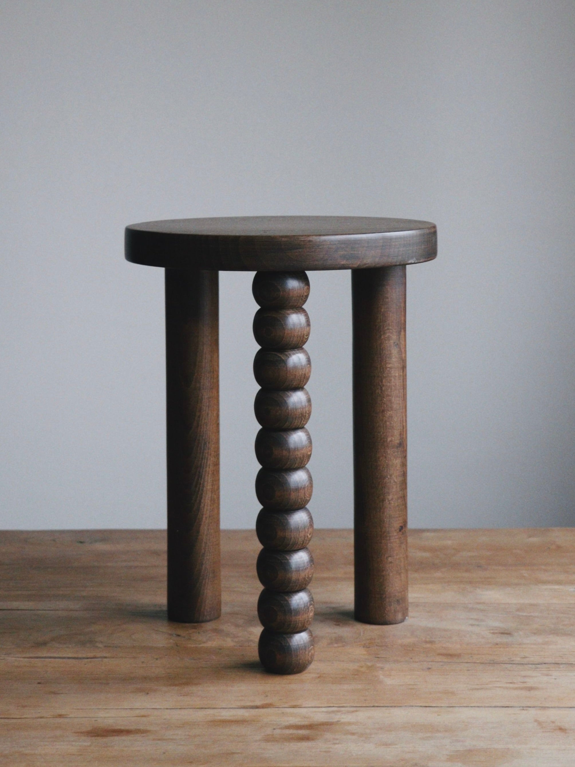 A wooden stool with three legs; two are smooth and cylindrical, while the third echoes the Piloti Side Table from Maitoinen Home, showcasing a unique design with Euclidean geometry comprised of stacked spherical segments. Its dark walnut finish enhances its elegance as it stands on a wooden surface against a gray background.