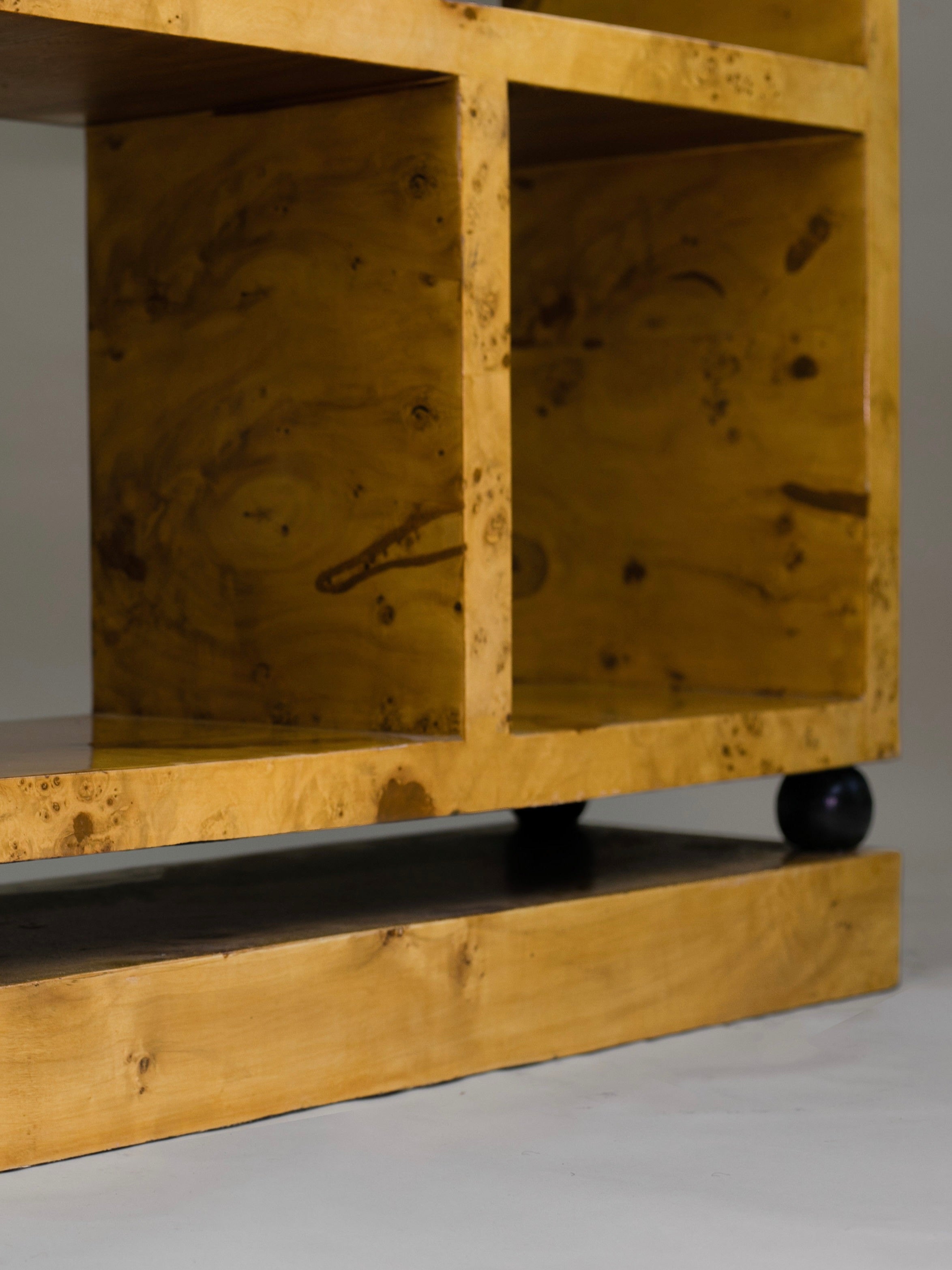 The Calisto Double Bookcase by Meola Interiors boasts a minimalist, mid-century design with its natural wood grain pattern, square compartments, small black wheels, and a smooth polished golden-brown finish.