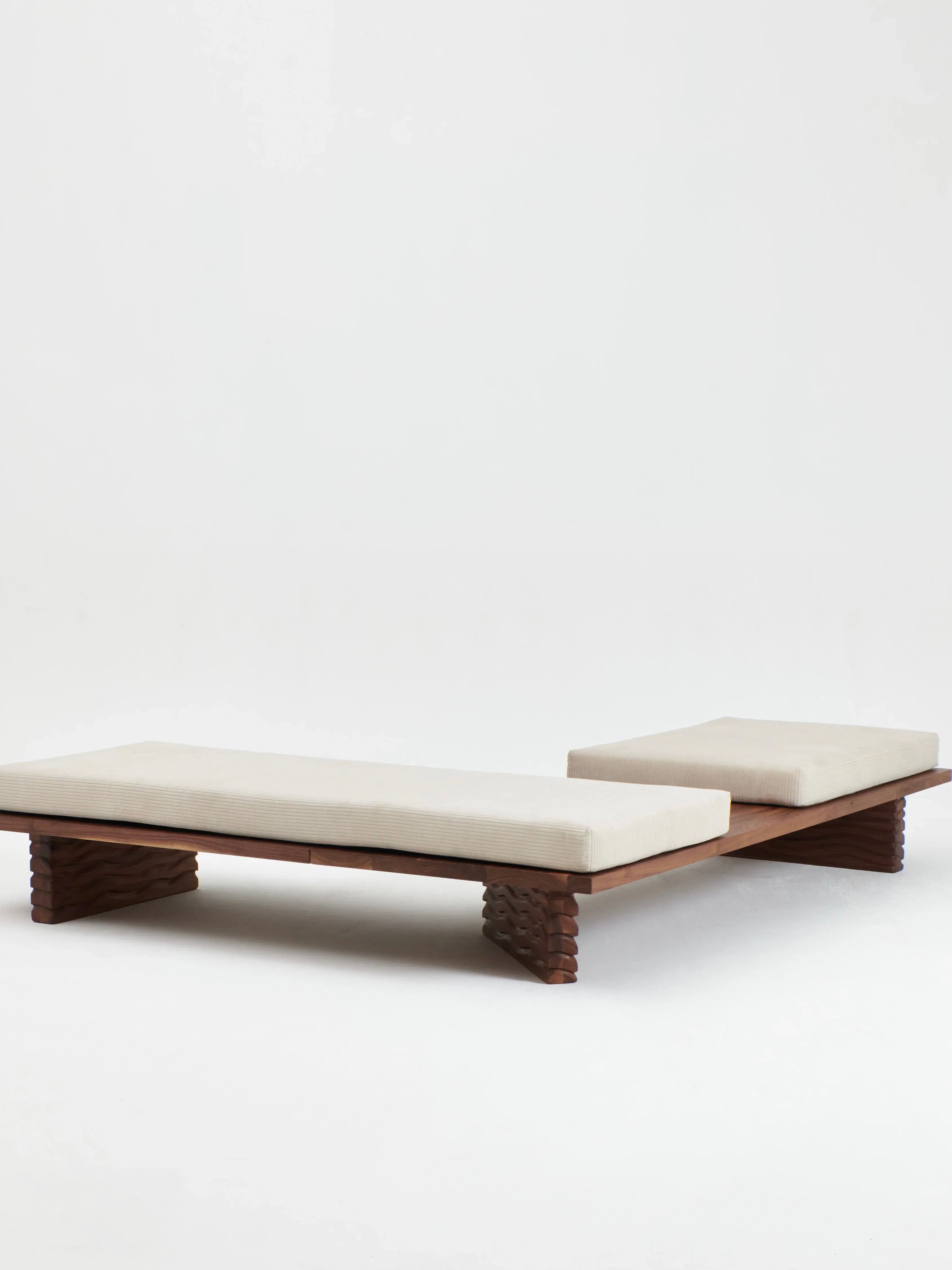 The Nazaré Daybed L Shape from Project 213A is a modern, minimalist daybed featuring a solid walnut frame with intricately carved legs and cushioned seats upholstered in light beige fabric. Handmade in Portugal, this low-profile design with clean lines offers a sleek and contemporary look.