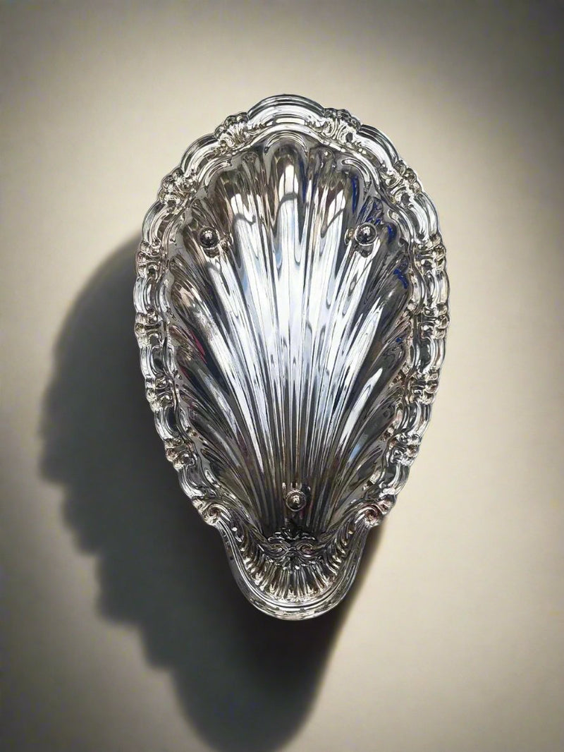 The Medium Shell Vide Poche by Les Objoies is a decorative, oval-shaped, silver-plated accessory resembling a seashell with intricate ridges and scalloped edges. It casts a soft shadow on a beige background, showcasing its ornately designed glossy, reflective surface that highlights its delicate details and meticulous craftsmanship.