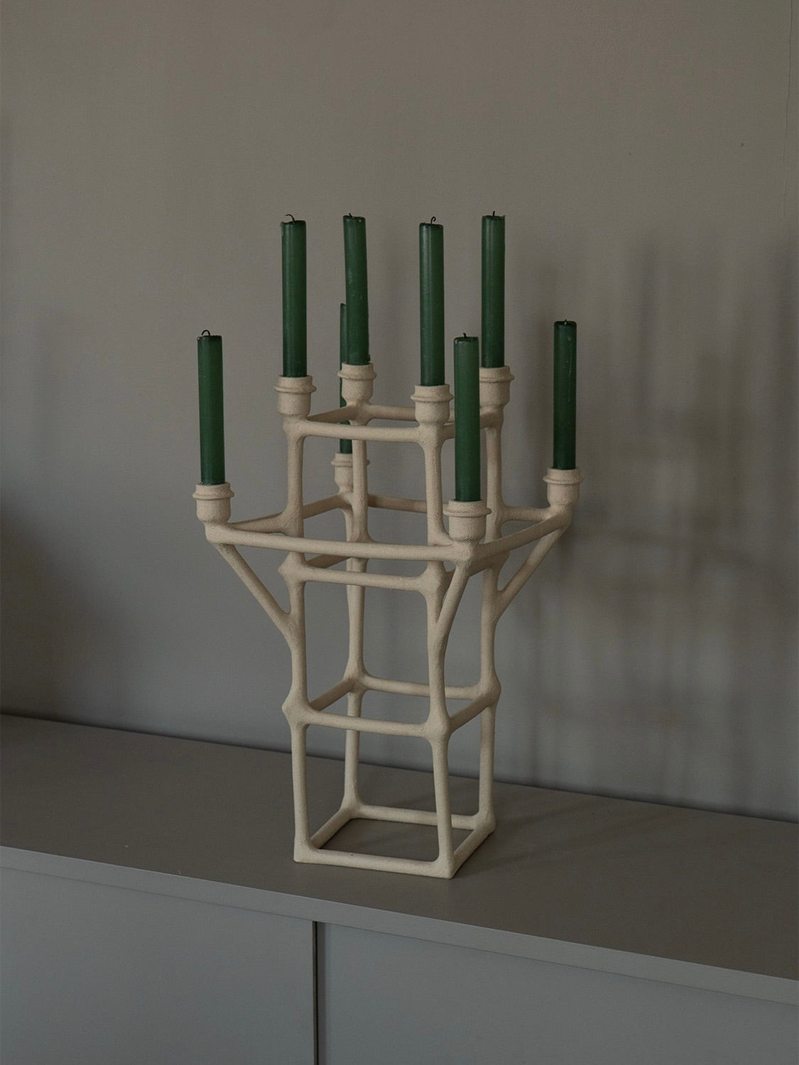 The "Clay Chandelier" by Solenne Belloir is a sculptural, handcrafted candle holder with a geometric flair, featuring multiple tiers that support nine green candles. This unique piece showcases an abstract, lattice-like design reminiscent of a chandelier, elegantly displayed on a gray surface against a plain gray background.