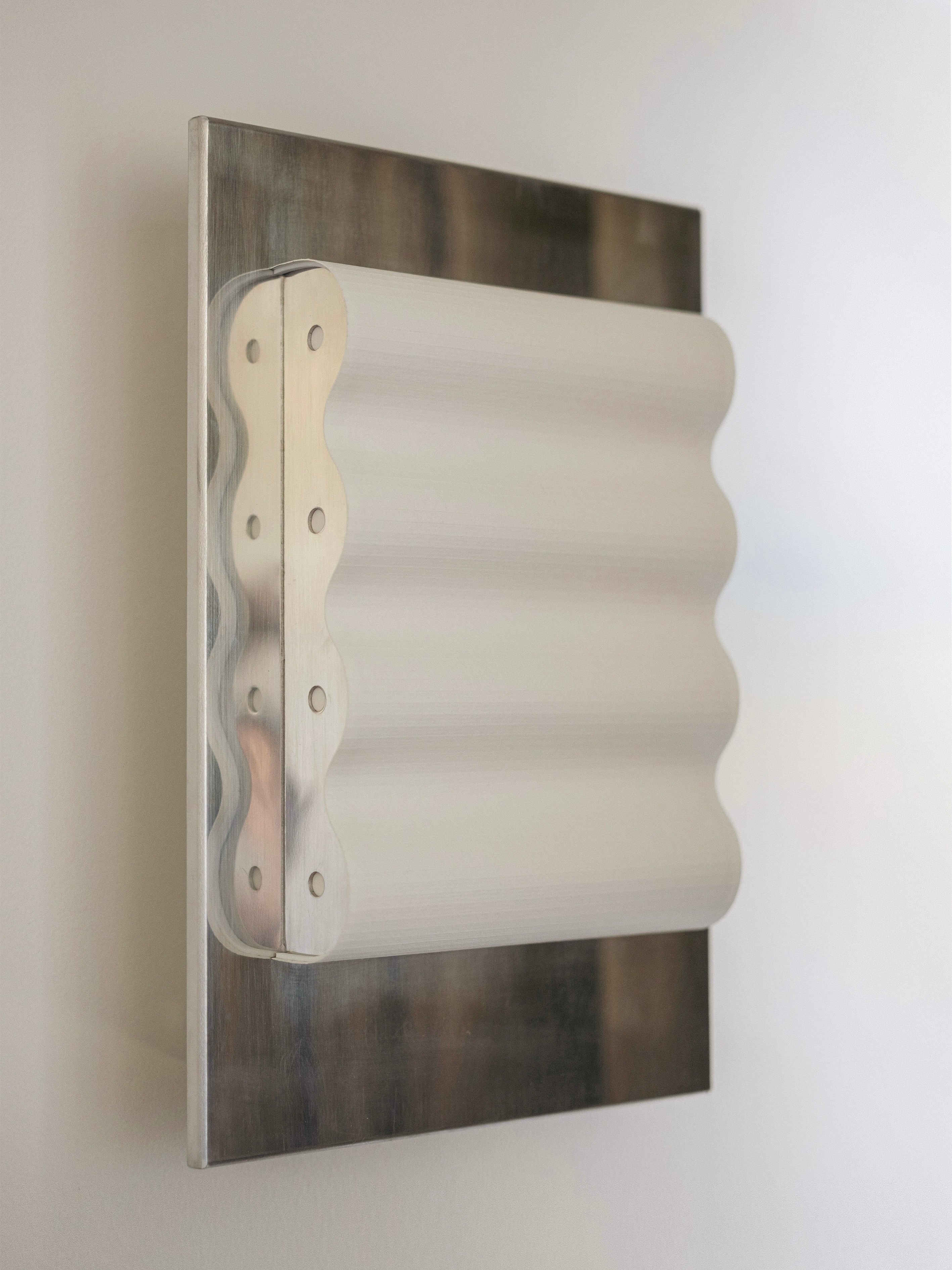 The Wall Light Frame Miroir M by Violaine d'Harcourt is a minimalist wall-mounted sculpture featuring a rectangular metal plate with a wavy, layered design protruding from its center. The artwork combines smooth, metallic textures with modern, abstract aesthetics akin to modernist architecture. The background is a plain, light-colored wall.