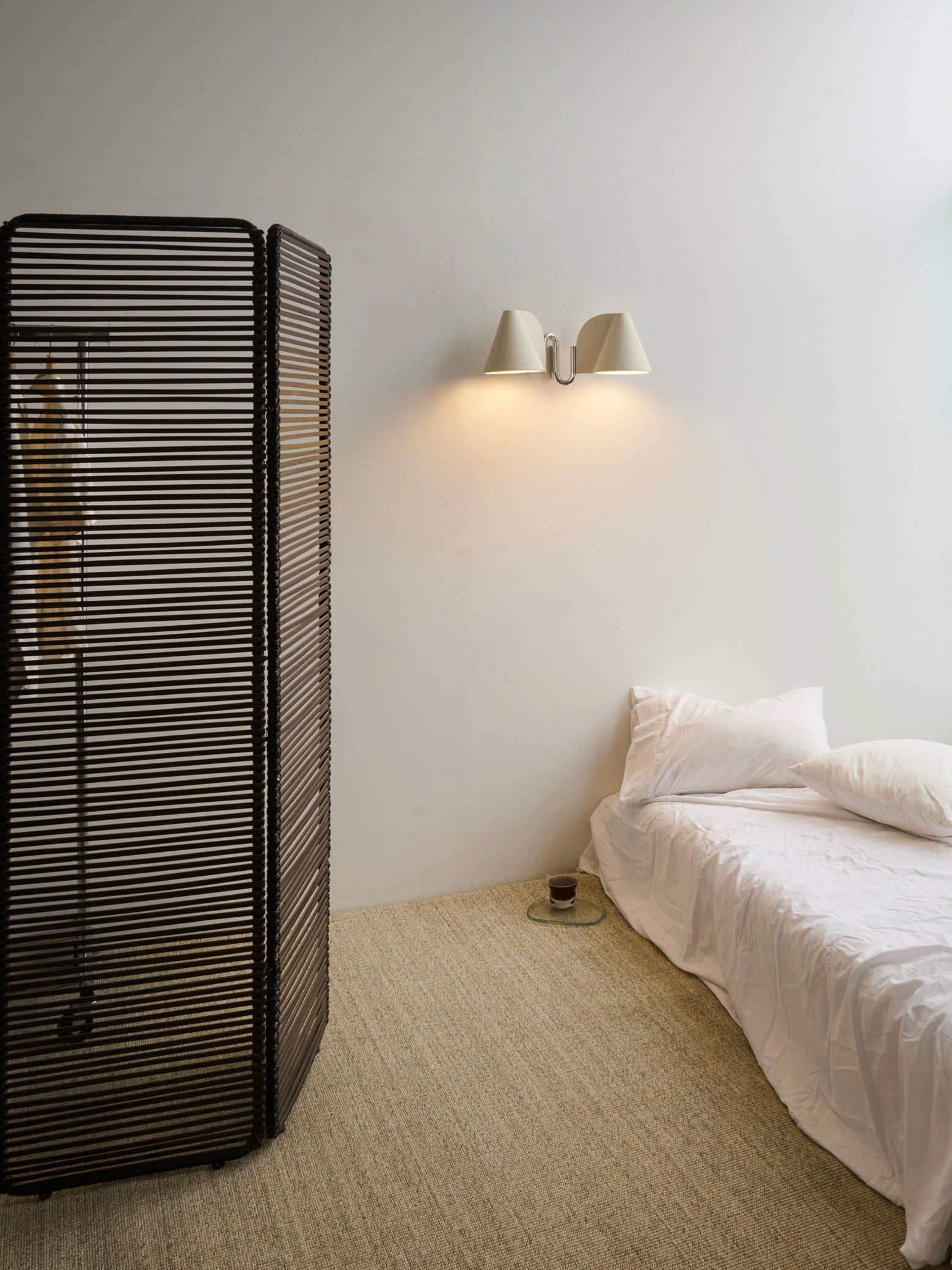 A minimalist bedroom showcases a single bed with white linens, a wooden room divider, and the 'Synapse' Wall Light by Mariza Galani casting a warm glow. A small round side table holds a candle against neutral-toned walls and carpet.