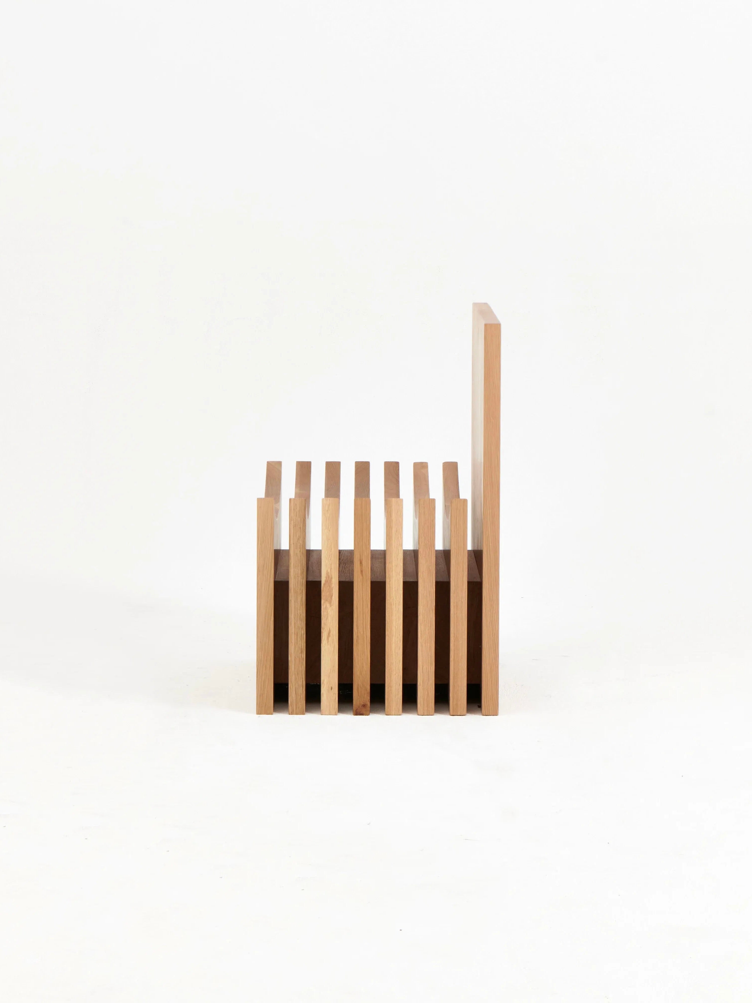 The Melides Chair by Project 213A is a minimalist, hand-crafted wooden chair characterized by vertically slatted sides and back, offering a modern, open design. Made of solid oak, its light-colored wood contributes to a clean, natural aesthetic. The chair’s simple structure underscores functionality and elegance against the plain white background.