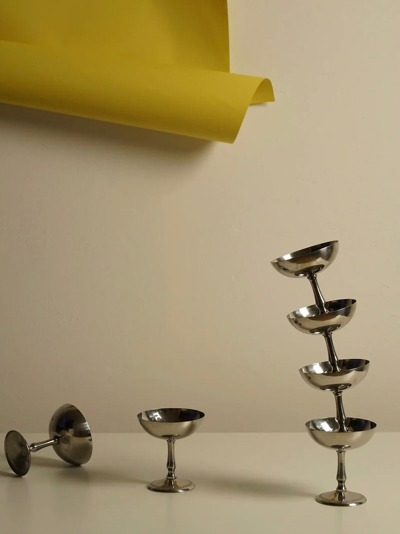 A minimalist scene features a tan background with a piece of yellow paper hanging from the top. On a white surface, four stainless steel ice cream cups from the Les Objets Amsterdam Set of 6 Ice Cream Cups are arranged: one lying on its side, one standing upright, and three stacked vertically to form an artful tower.