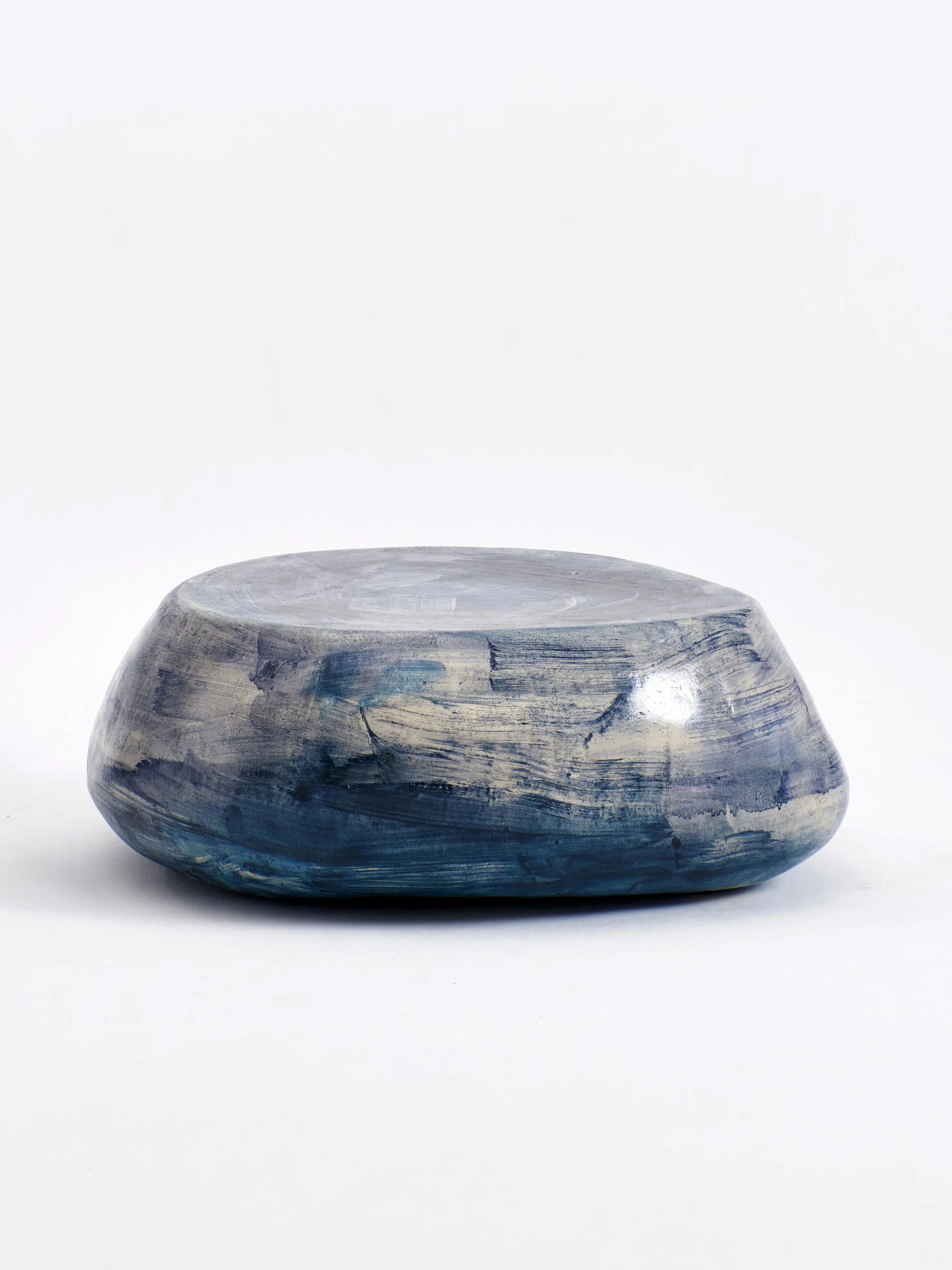 The Brushed Blue Ceramic Coffee Table by Project 213A is a low, round piece designed with a smooth, stone-like appearance and handmade craftsmanship. Its surface blends gray and blue hues, creating a weathered, marbled effect against a plain white backdrop.