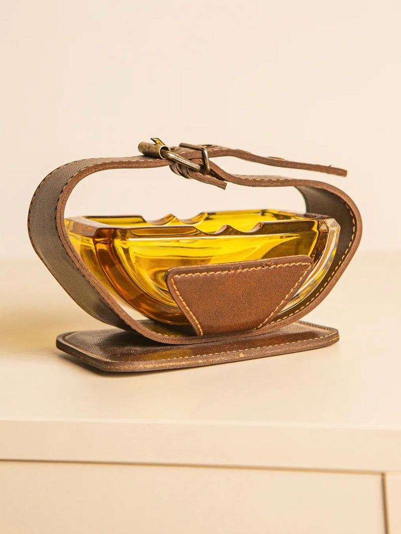 The Italian Ashtray from the 60s by Treaptyque features a distinctive, oval-shaped amber glass bowl nestled in a decorative brown leather holder with a handle and buckle that brings forth a vintage aesthetic. This artistic leather wrap envelops the bowl, reminiscent of an Italian ashtray, beautifully displayed on a light-colored surface.