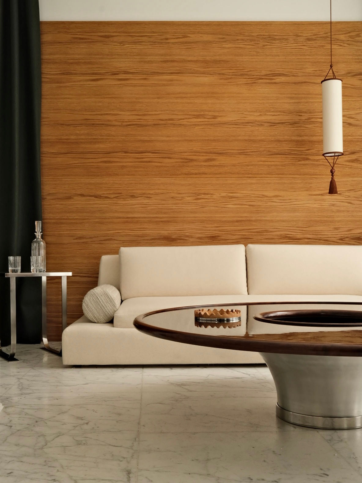 A modern living room showcases the "Podium" sofa by HAUVETTE & MADANI, inspired by seventies design in a beige hue, set against a wooden accent wall. A round wooden table rests on a marble floor. A glass decanter and two glasses are positioned on a small side table, while a cylindrical pendant light is suspended from above.