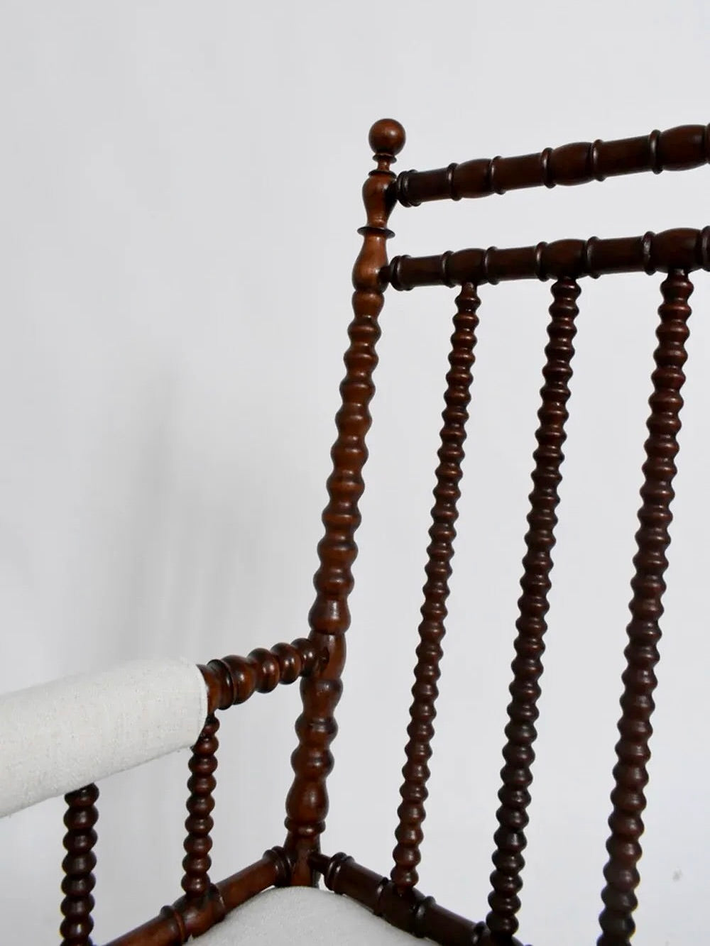 Close-up of a Barracuda Interiors Pair of Wood Armchairs, 19th Century, featuring a spindle backrest and armrest. These chairs, evocative of 19th Century England, boast a polished finish and white upholstered seats. The evenly spaced spindles enhance the chairs' traditional and elegant design against a plain background.