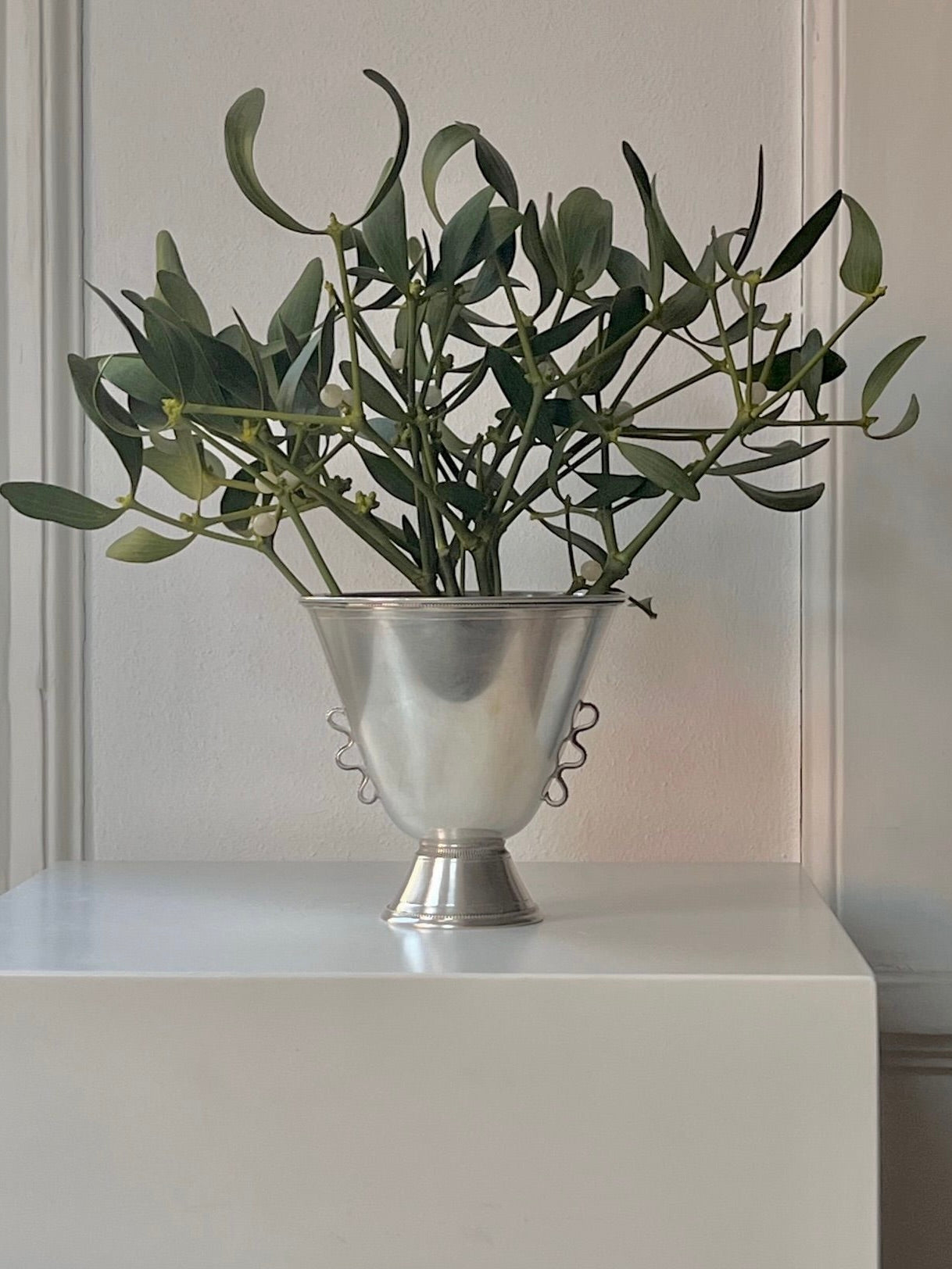 The 1930s Silver Vase by Médecine, featuring intricate craftsmanship and elegant handles, holds a green plant atop a white pedestal against a light gray wall, evoking the simple yet sophisticated aesthetic of Stockholm in the 1930s.