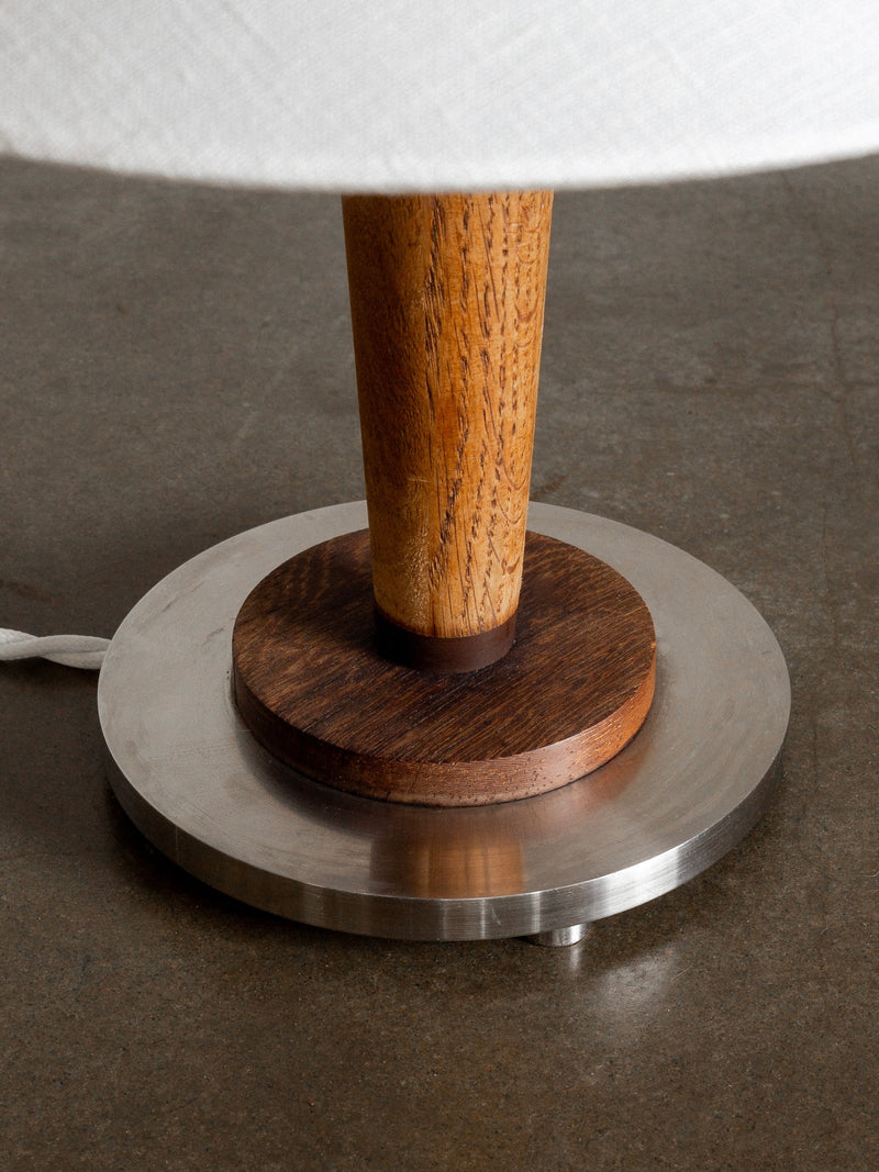 Close-up of the Spigel Mid-Century Metal and Wood Lamp. This Art Deco-style lamp features a wooden stem connecting to a round metal base accented with teak wood. The textured wood creates a striking contrast against the smooth metal surface. A white cord extends from the back of the base, and the lamp is situated on a dark, polished surface.