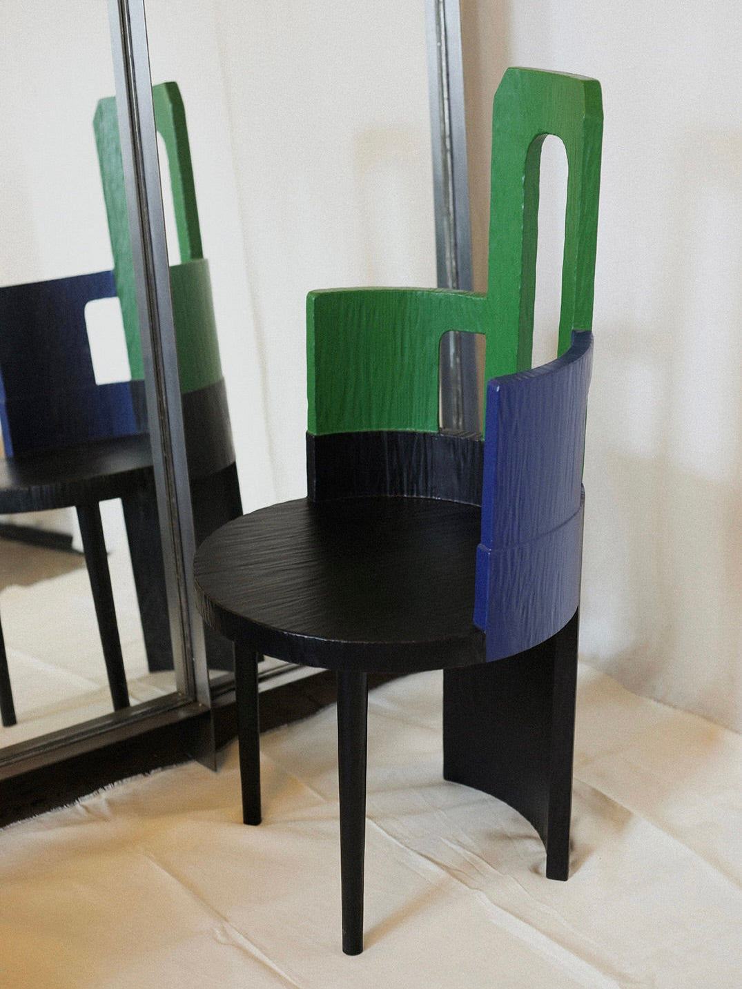 The Sculptural Chair III in Primary Colors by Rooms Studio has a round black seat and three backrest segments in green, blue, and black. Its striking design is mirrored on a white surface, showcasing an artful blend of primary colors.