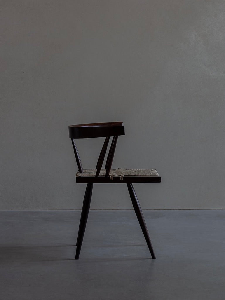 The "Manner of Nakashima - Grass Seated Chair" by Bicci de' Medici features a minimalist wooden design with a woven seat against a grey backdrop. Its dark rosewood finish and clean lines offer a classic and modern look reminiscent of George Nakashima's mid-century style.