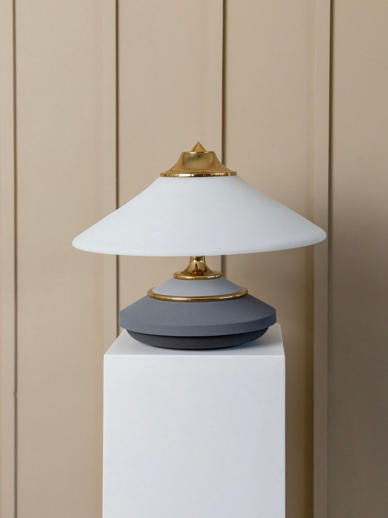 The Bottega Jacobs Memphis Style Vintage Murano Glass Table Lamp is a stylish piece, featuring a geometric design echoing the 1980s Memphis aesthetic. It boasts a wide white conical shade with gold accents atop a gray base, elegantly perched on a white pedestal against a beige paneled wall.