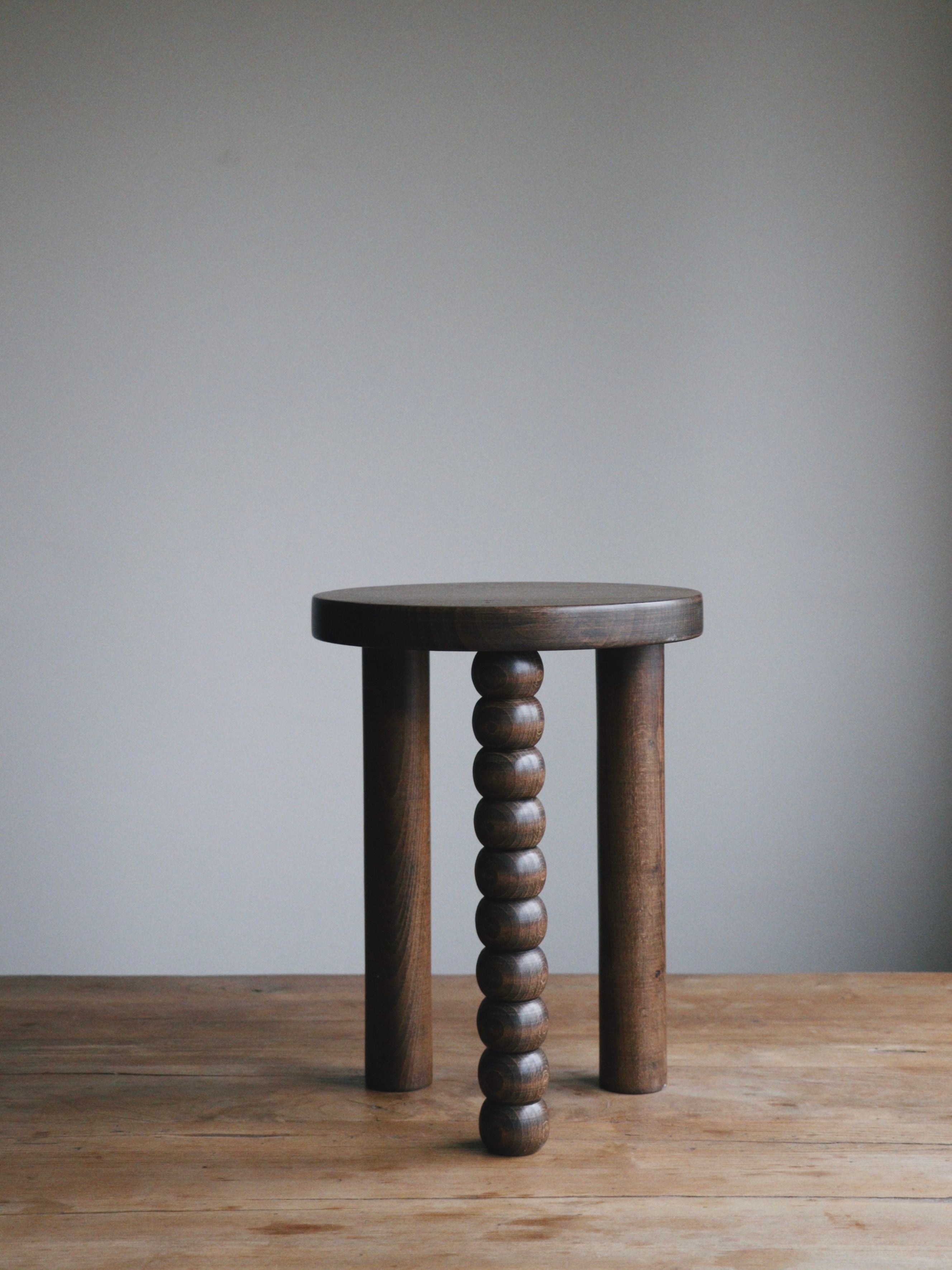 A uniquely designed wooden stool by Maitoinen Home, inspired by the Piloti Side Table, showcases three straight legs and one intricately carved with round, stacked shapes. Its dark walnut finish elevates its elegance as it rests on a wooden surface against a simple backdrop.