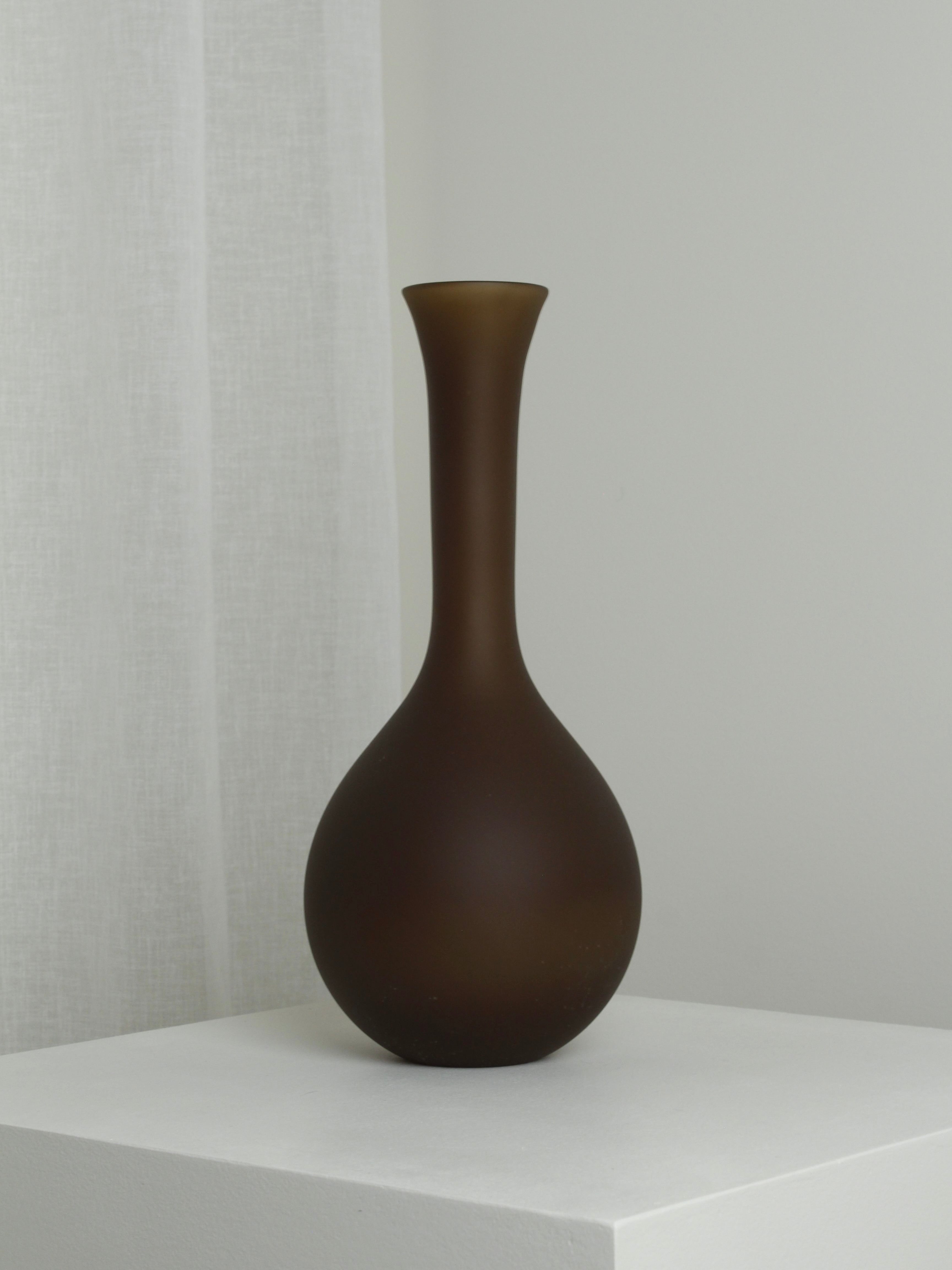 The XLarge Vase in Chocolate by Verre d'Onge rests elegantly on a white pedestal. Featuring a chocolate-brown hue and matte finish, this mouth-blown vase showcases a narrow neck that subtly flares at the top, paired with a rounded base for an elegant and minimalist design. A white curtain can be seen partially in the background.