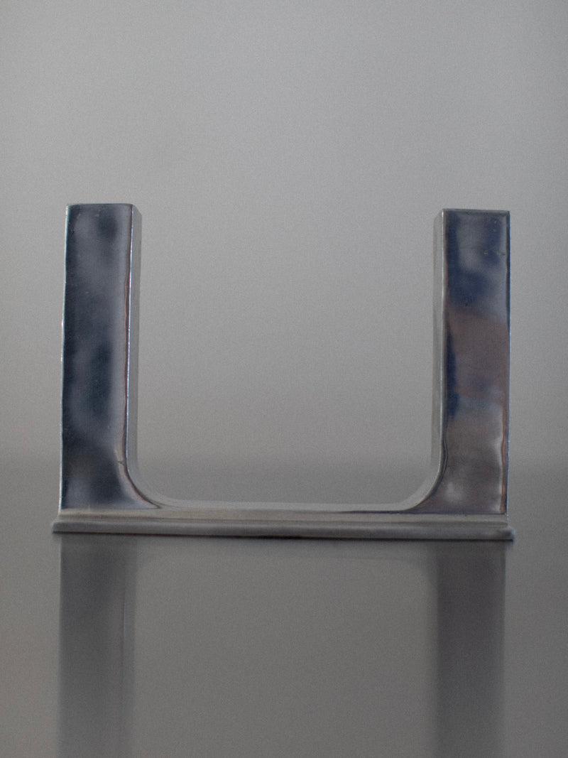 A minimalist Chrome Candleholder by Out For Lunch, resembling a sleek steel candleholder, stands on a reflective surface against a plain gray background. The candleholder has a smooth, polished finish and appears to be made of metal.
