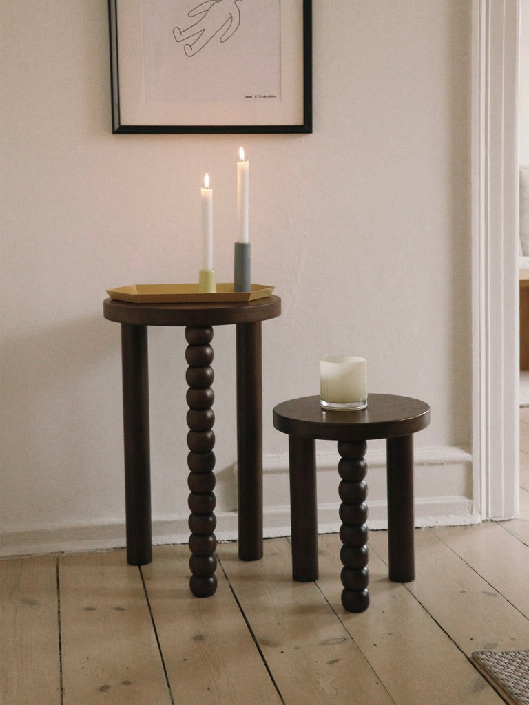 Two Piloti Side Tables from Maitoinen Home, featuring bead-like legs and crafted from light wood, stand by a white wall with abstract art. The taller table holds three lit candles, while the shorter one displays an unlit candle in a glass holder, subtly hinting at Euclidean geometry.