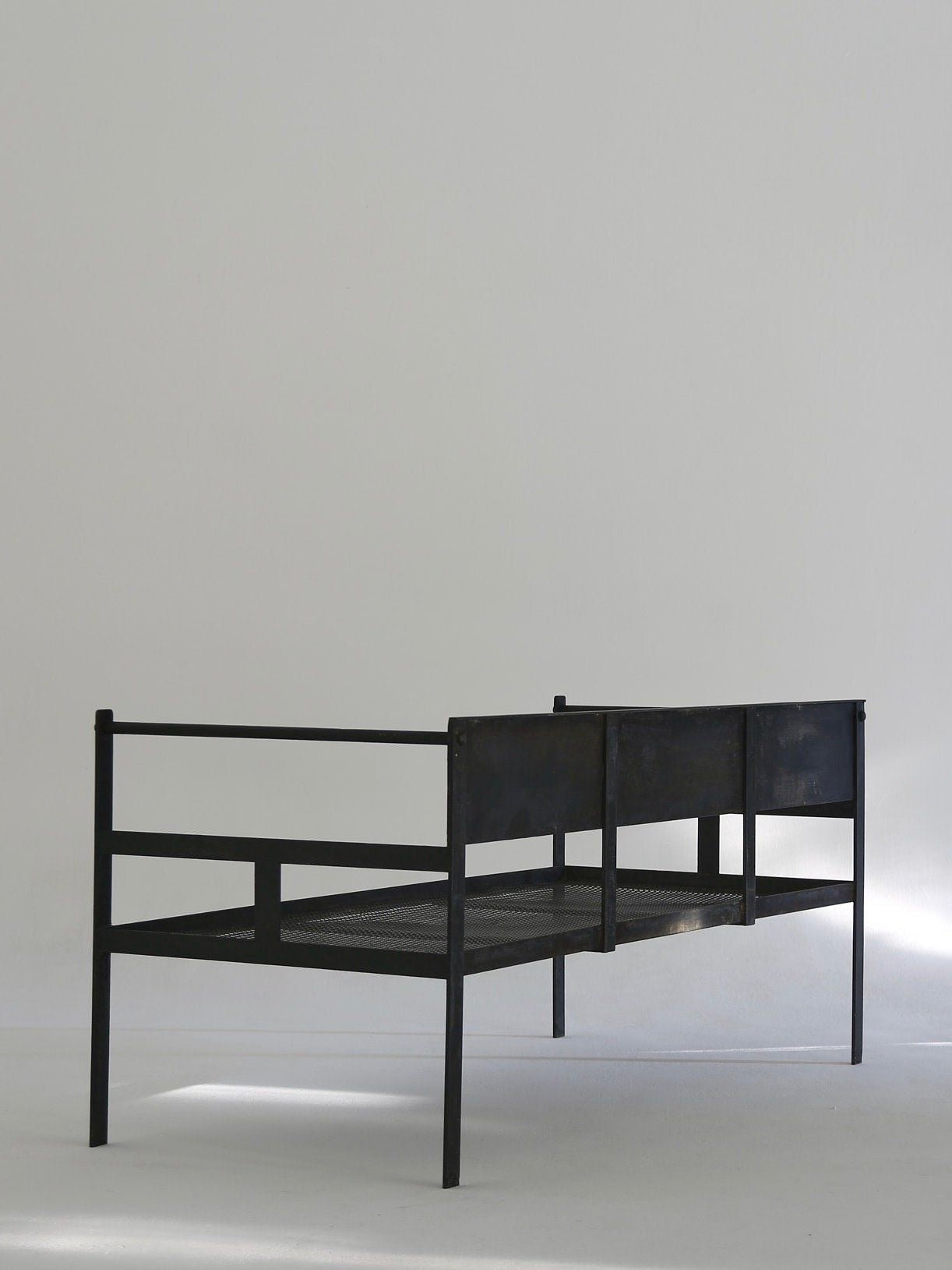 The Ancien et Jolie Modernist Lounge Set, a minimalist black metal design, complements a bright room with plain white walls. With simple vertical and horizontal bars and a low headboard, it casts soft shadows on the floor, reminiscent of Mathieu Mategot's design ethos.