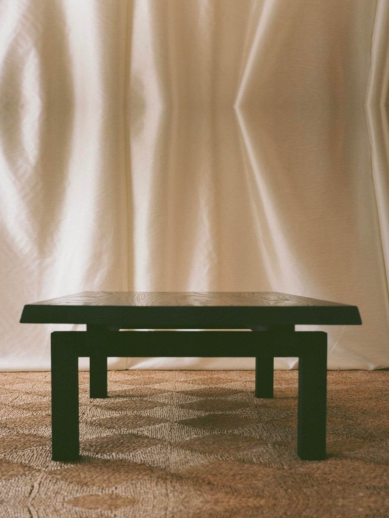 The Lantern Coffee Table by SWM STUDIO—a low, dark wooden piece echoing Japanese aesthetics—rests on a textured brown carpet before softly draped, light-colored curtains, enhancing the serene atmosphere with its minimalist solid oak design.