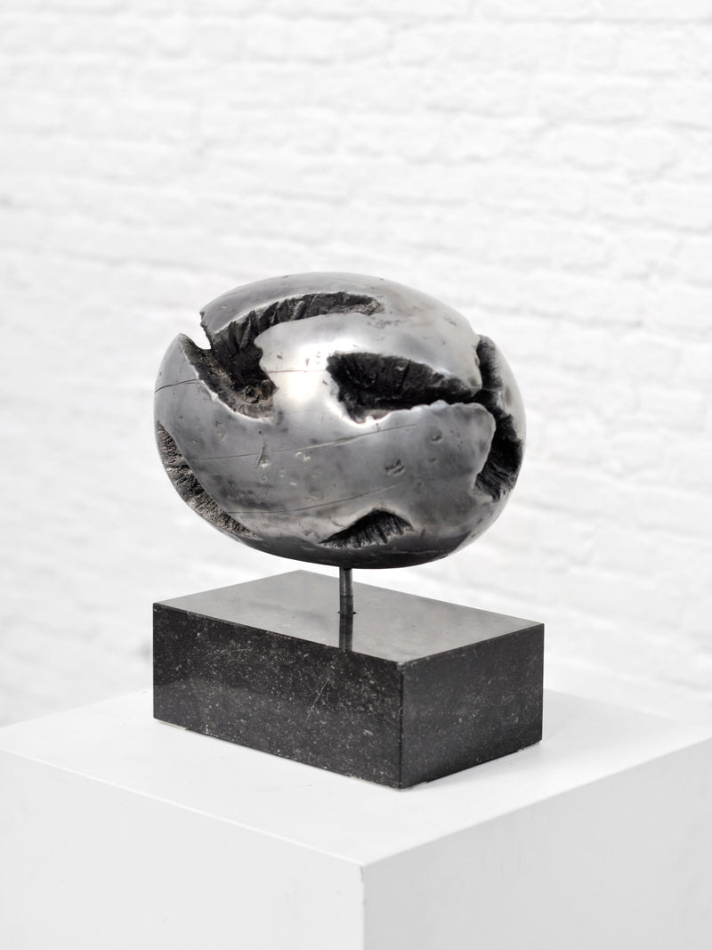 The 1970's Brutalist Spherical Metal Sculpture by Introverso is a striking modern art piece with a metallic, abstract spherical shape featuring rough textures and deep grooves. It rests on a black rectangular base, displayed on a white pedestal against a pristine white brick wall.