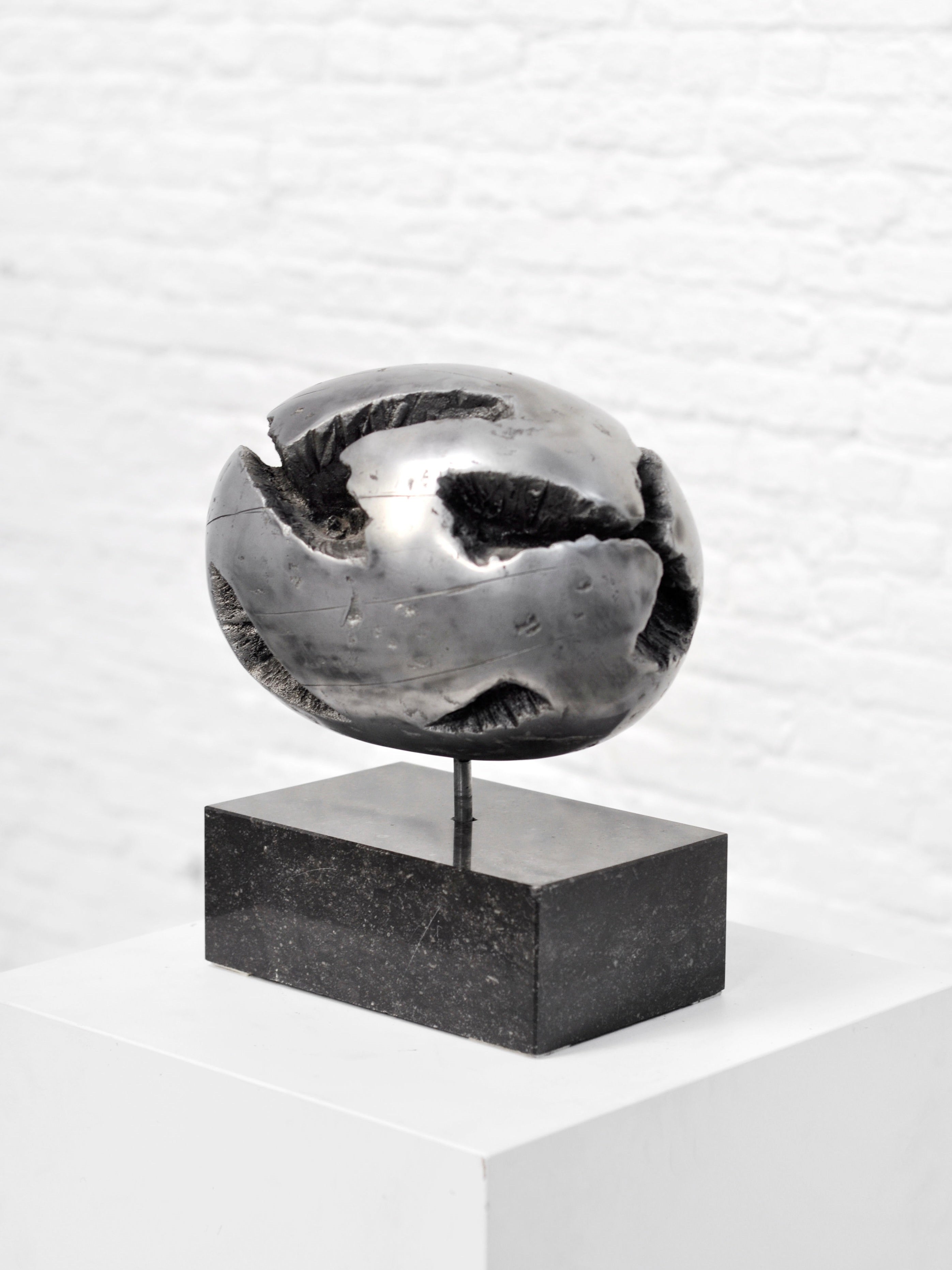 The 1970's Brutalist Spherical Metal Sculpture by Introverso is a striking modern art piece with a metallic, abstract spherical shape featuring rough textures and deep grooves. It rests on a black rectangular base, displayed on a white pedestal against a pristine white brick wall.