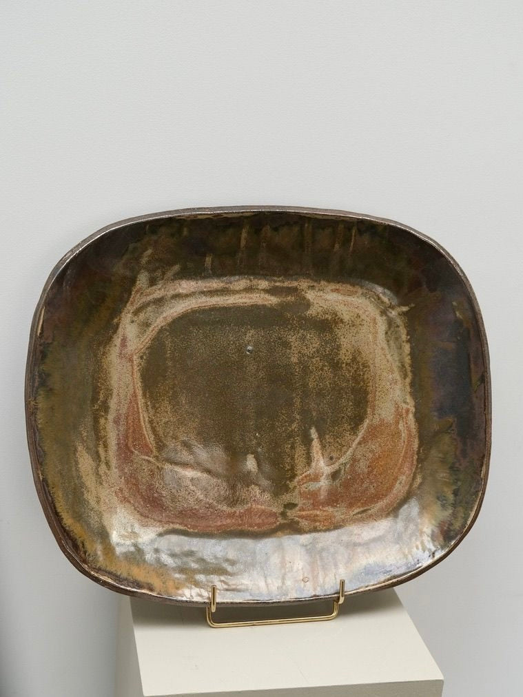 A square, glazed stoneware dish from LA NOUVELLE GALERIE rests on a metal display stand. Reminiscent of Pierre Culot's work, this Large Decorative Stoneware Dish features an earthy, mottled glaze in shades of brown, green, and beige with a slightly uneven surface. The stand is placed on a light-colored pedestal against a plain background.