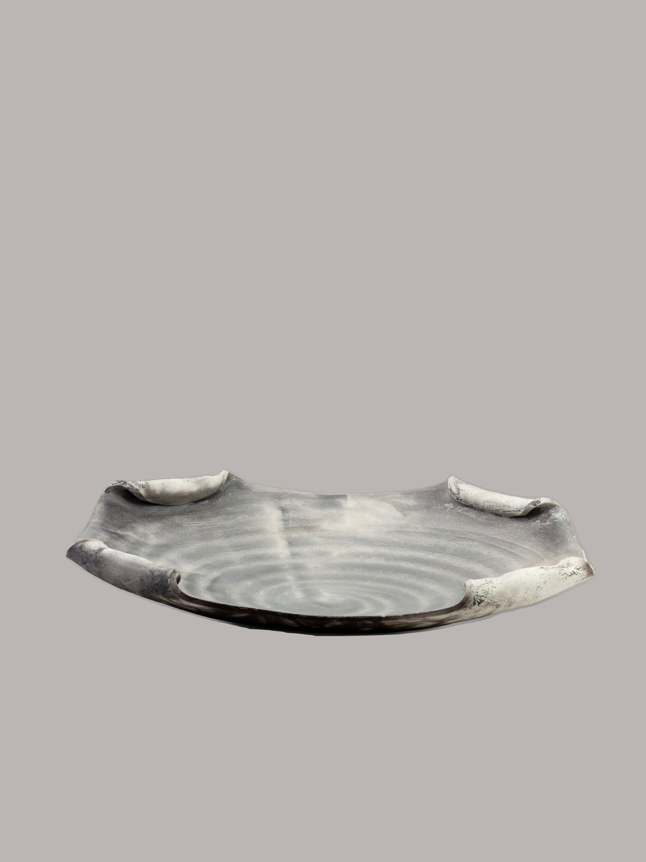 A shallow, round, dark grey ceramic dish with a matte finish sits against a plain grey background. This Large Raku Platter by RELIC LONDON features subtle, irregular edge protrusions and a smooth spiral pattern at the center. Its handcrafted appearance makes it an exquisite centerpiece.