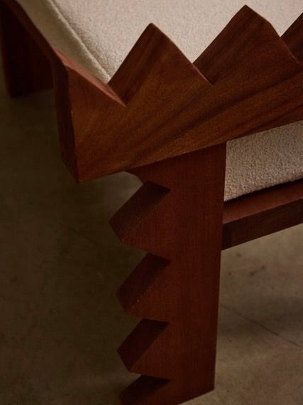 Close-up of the Jura Bench by Ah Um Design Studio, featuring unique zigzag edges on the backrest and legs. The hand-carved wood seat is upholstered with light-colored fabric, contrasting beautifully with the dark solid mahogany. The design showcases intricate craftsmanship and distinctive geometric detailing.