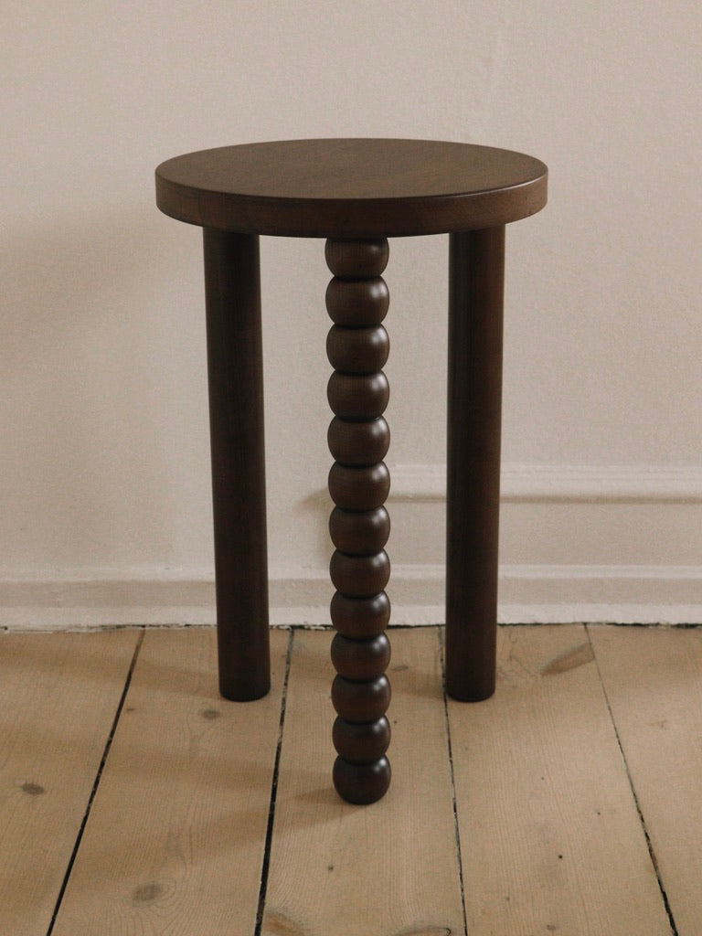 The Piloti Side Table by Maitoinen Home is a minimalist wooden piece in dark walnut with a round seat and three legs on a wooden floor, featuring one uniquely designed leg with stacked bead-like shapes, echoing Euclidean geometry for a simple, modern look.