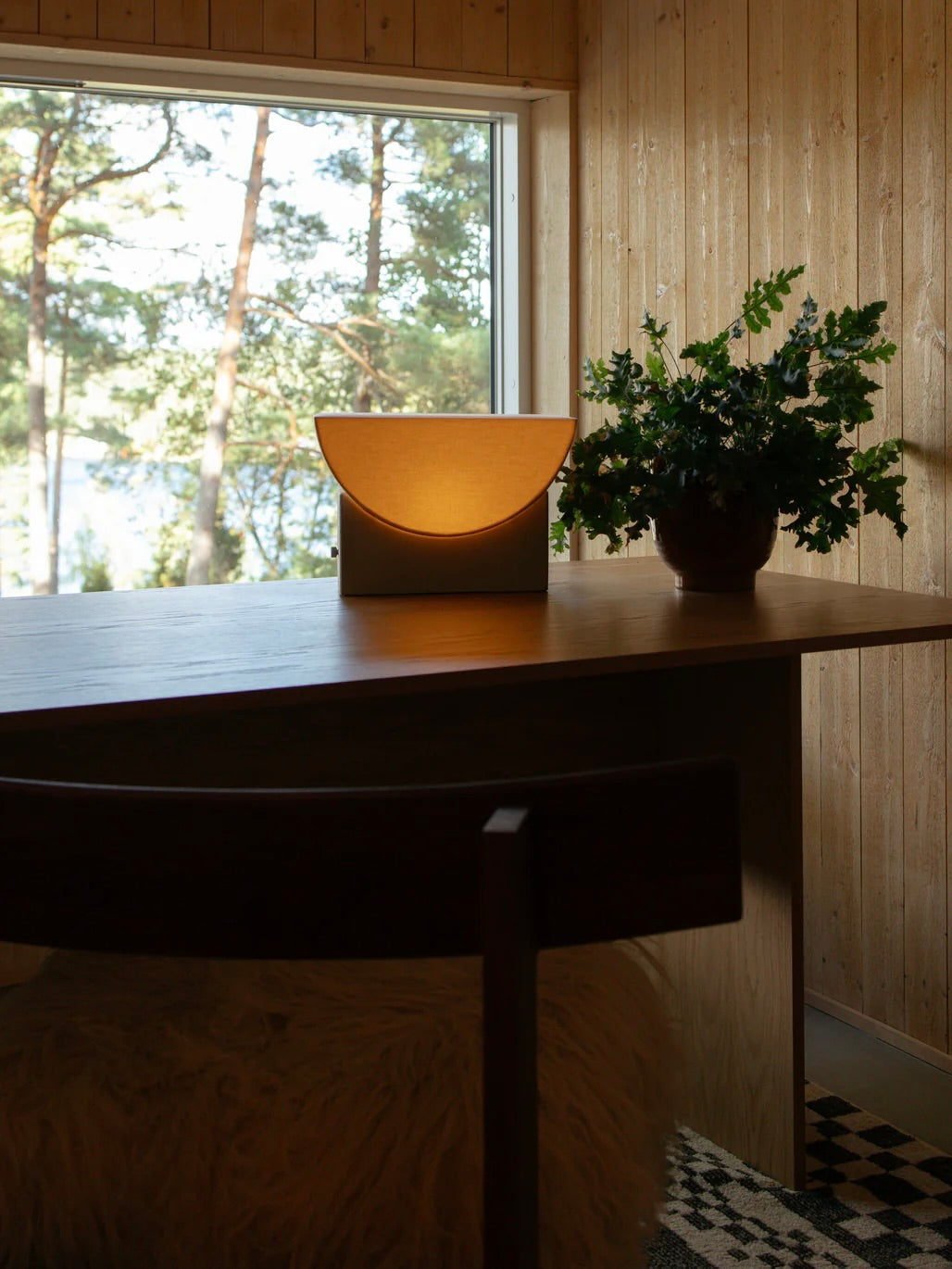 A cozy wooden room with a large window overlooking pine trees and a lake features a wooden table displaying the "Reminder" Table Lamp by Enkei Lights AB, along with a vase filled with green foliage accentuated by geometric forms. A chair with a fluffy seat is partially visible, bathed in natural light.