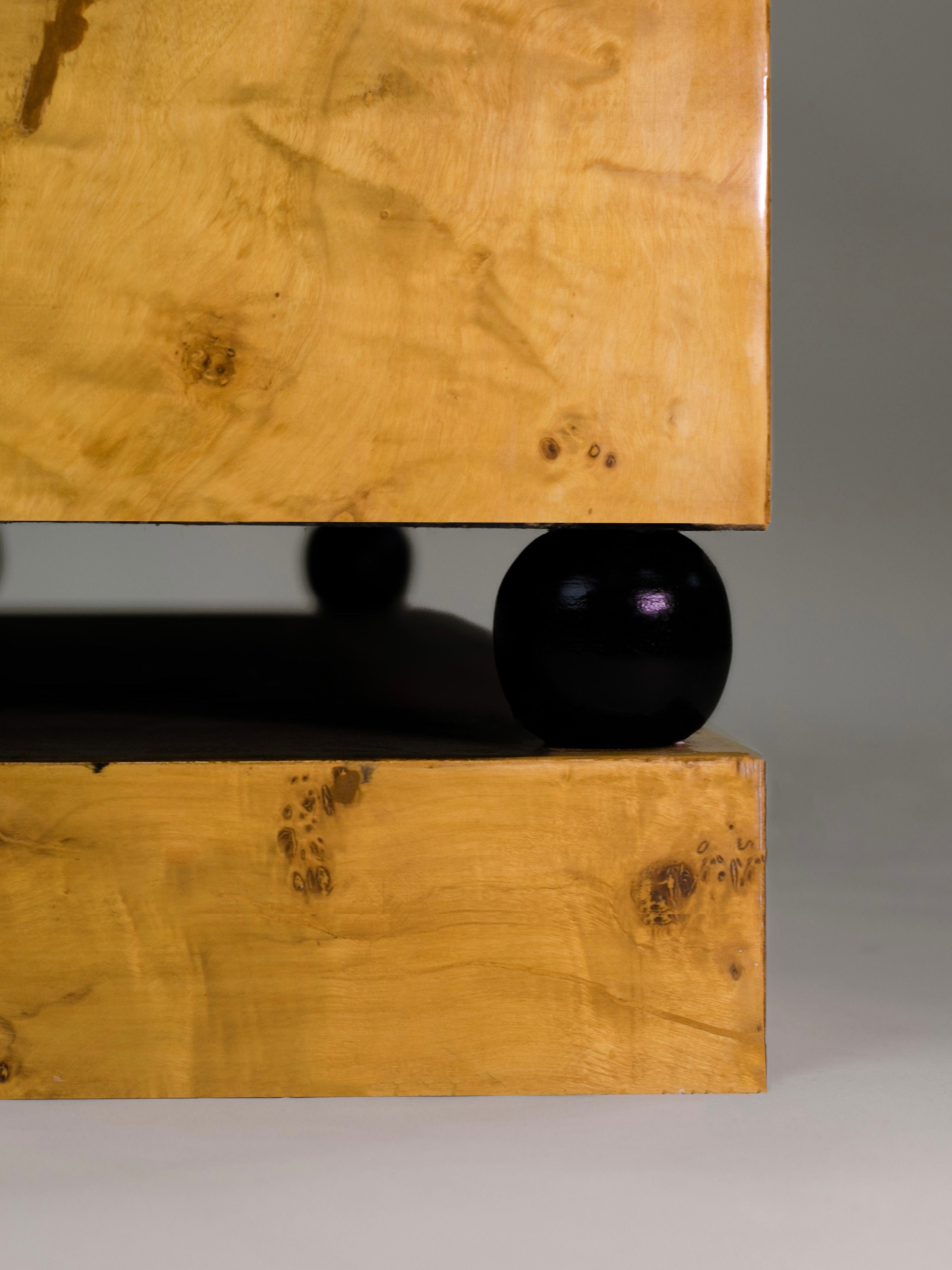 The 'Calisto' Double Bookcase by Meola Interiors features a minimalist design with a polished finish. A dark spherical element is nested between two rectangular slabs, showcasing visible wood grain and natural knots for a mid-century look against a soft gray background.