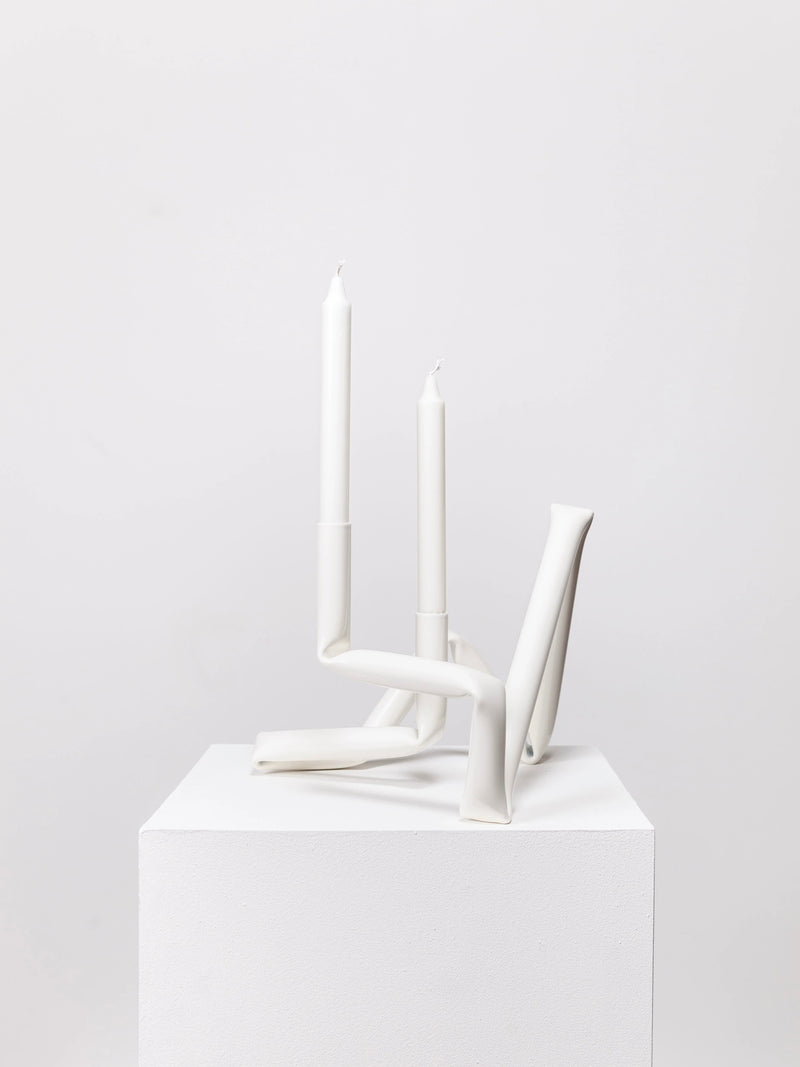 The Bucatini candle holder (White) from AOT STUDIO's Primi collection features a modern, minimalist design with an abstract, twisted form that holds two tall, unlit white candles. Hand-crafted and elegant, this piece is showcased on a white pedestal against a plain light gray background.
